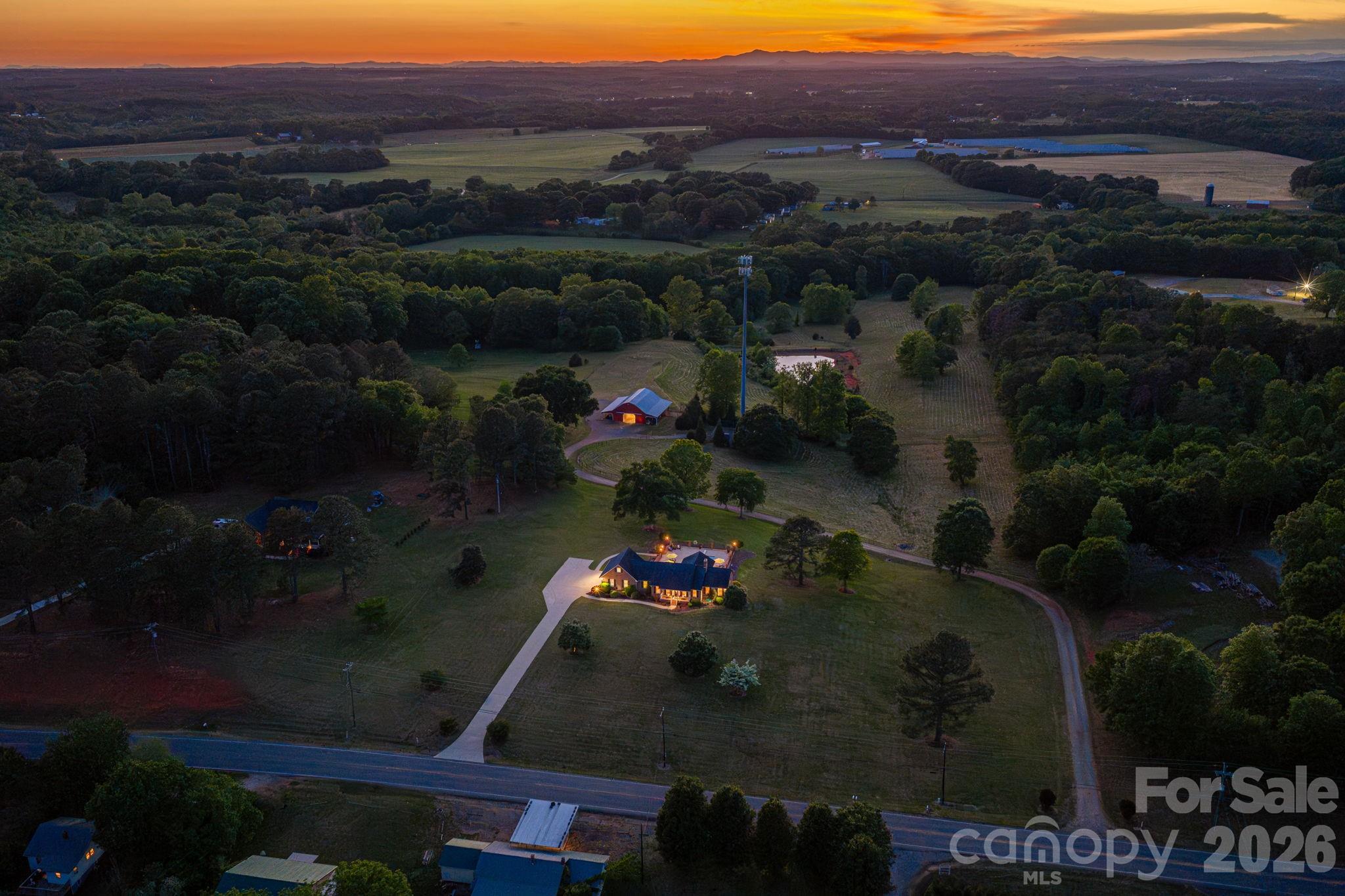 Aerial view of a home and wooded acreage at sunset, with a long driveway and farmland.