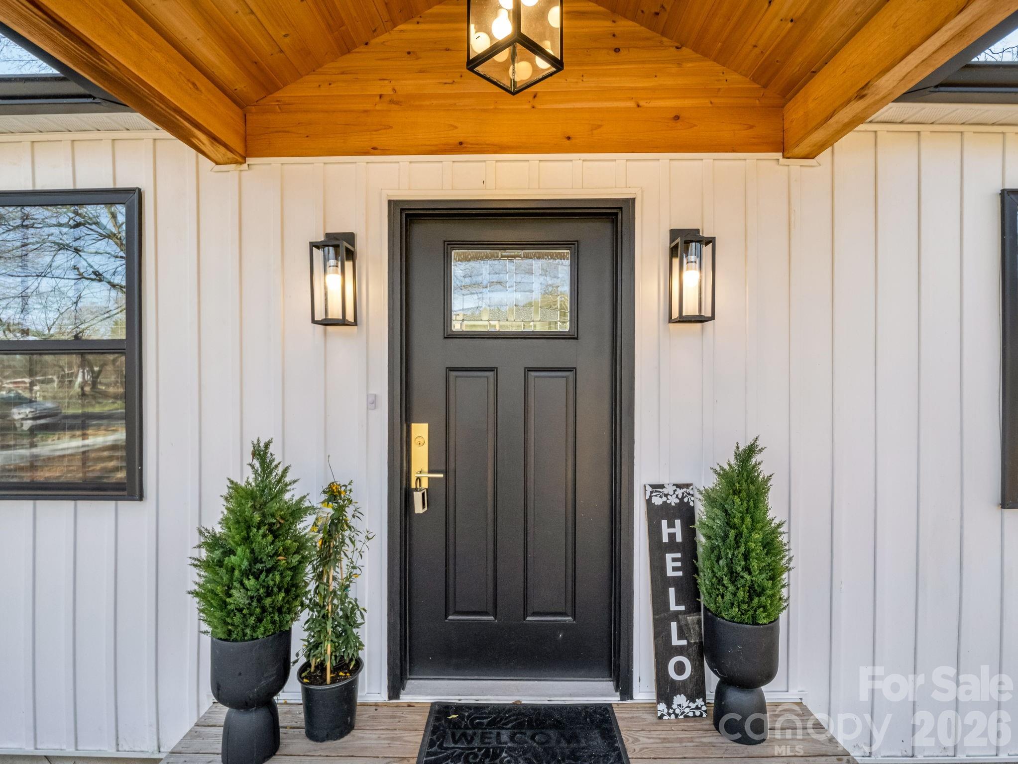 Covered front porch with a black entry door, warm wood ceiling, matching lanterns, and potted evergreens.