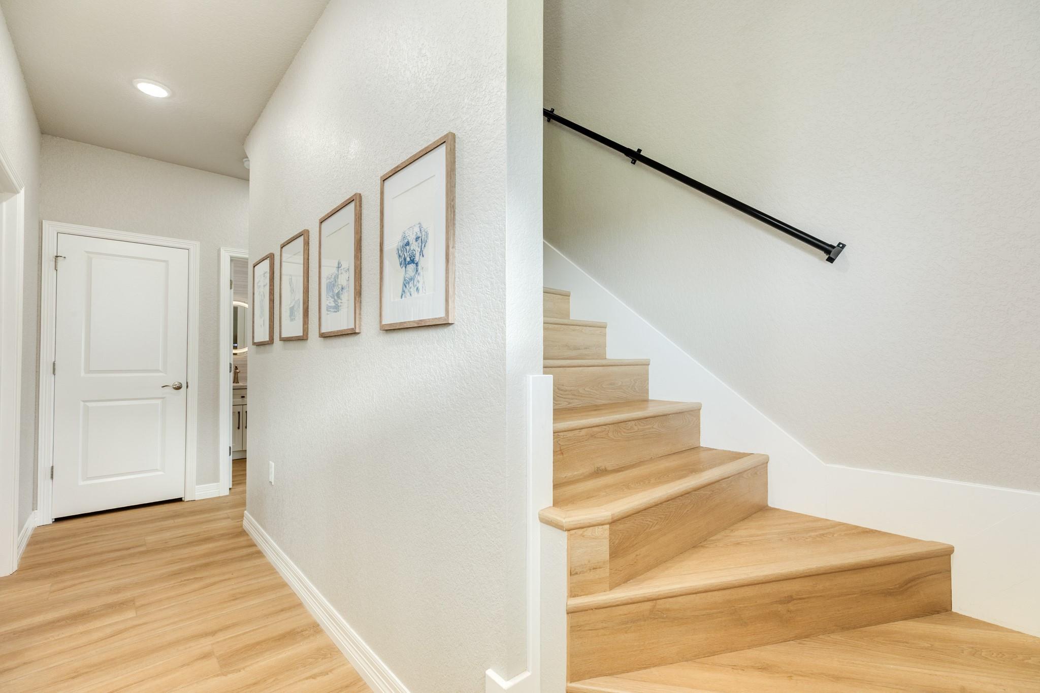 Bright hallway with light wood floors, framed prints, and a staircase with a black handrail.