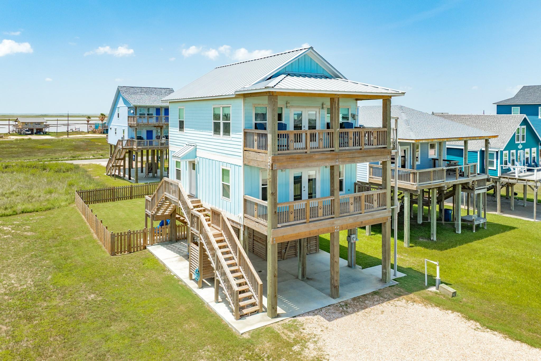Elevated light-blue coastal home with broad wooden balconies, exterior stairs, and water in the distance.