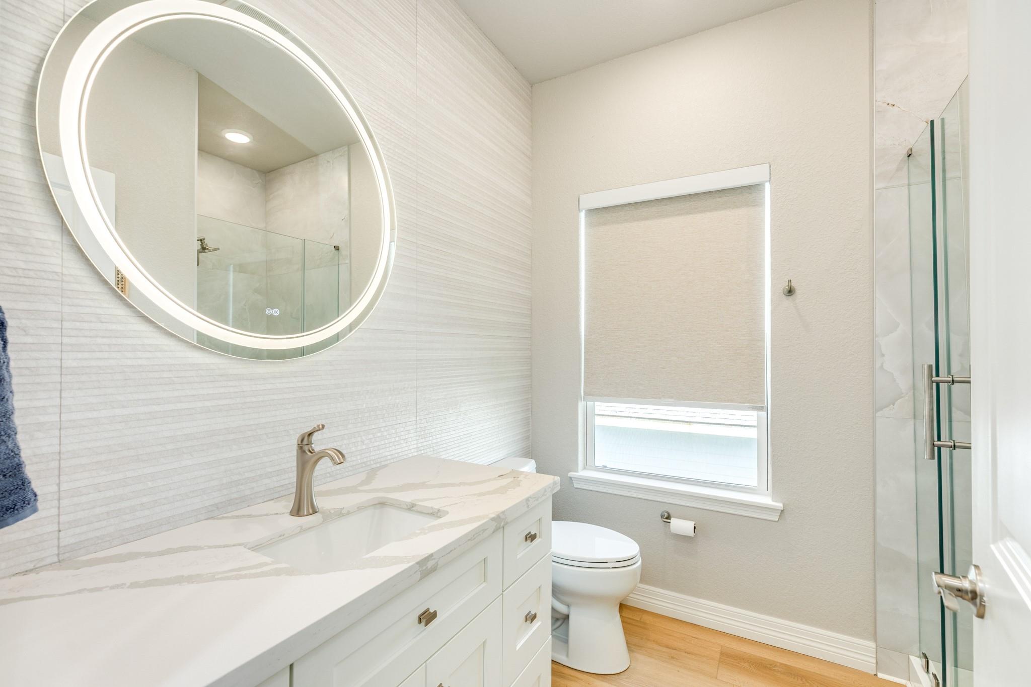 Modern bathroom with a marble-look vanity, illuminated round mirror, shaded window, and glass shower.