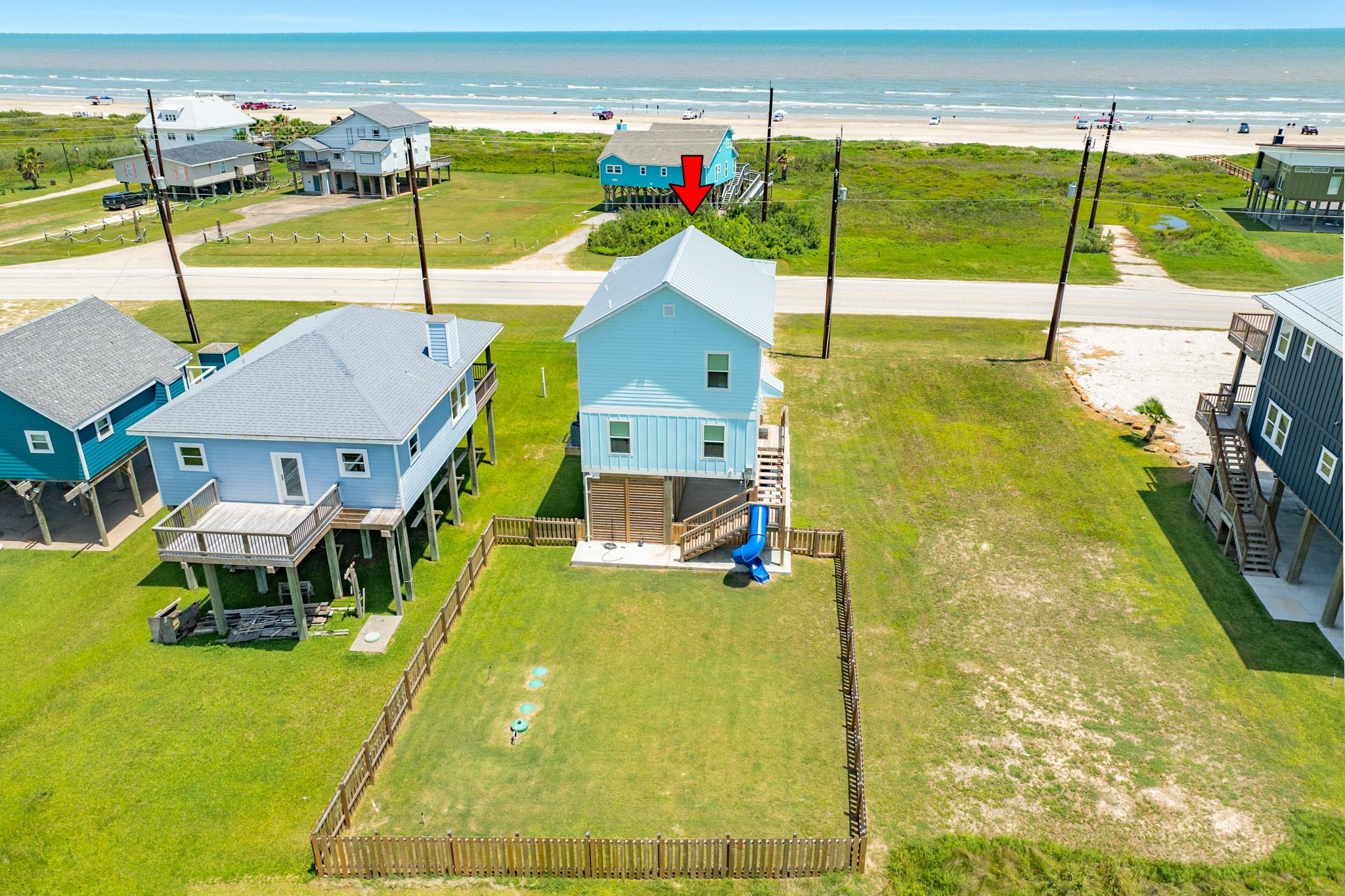 Aerial view of a blue coastal house and fenced yard, with the beach and ocean beyond.