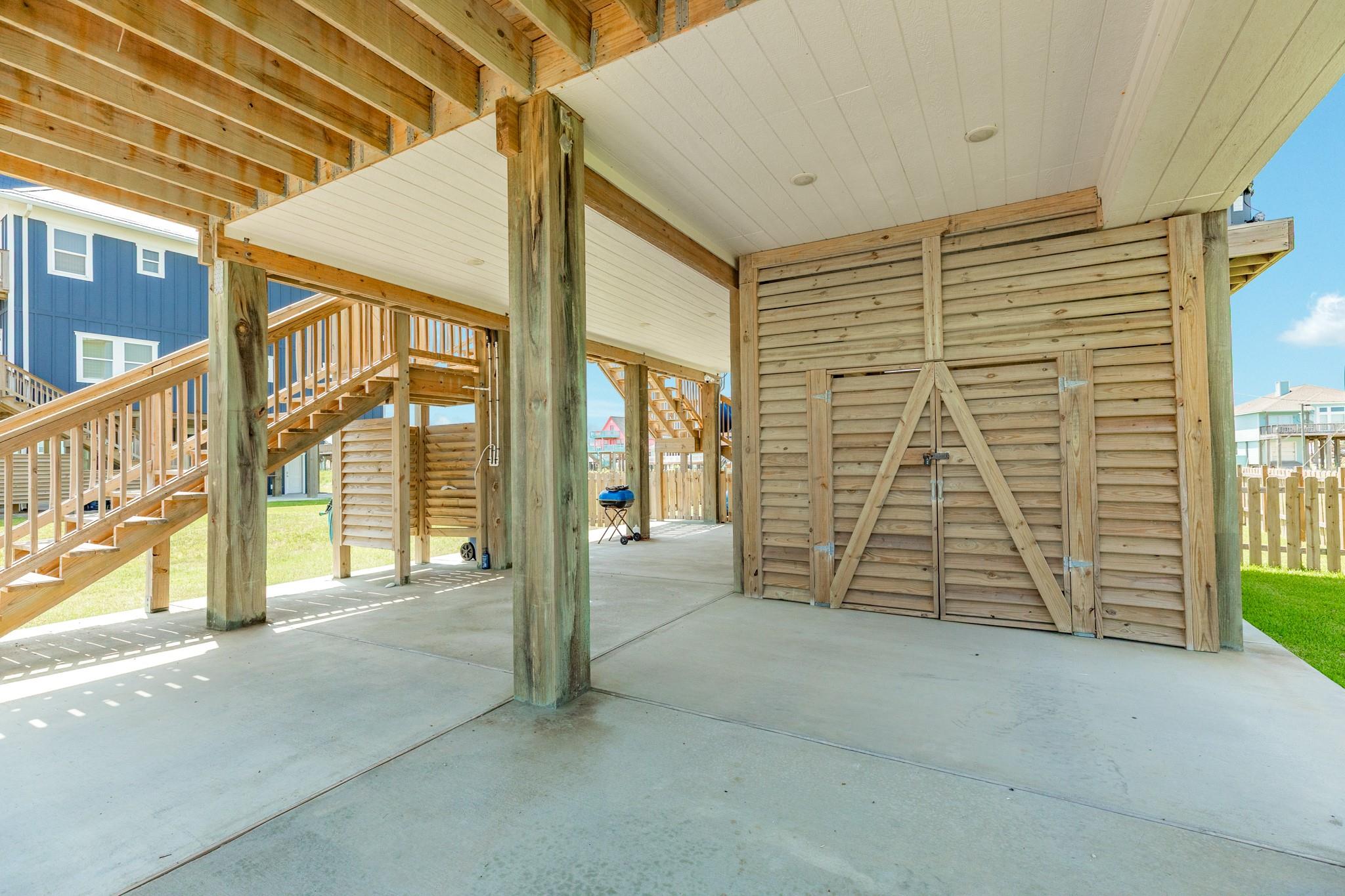 Covered area beneath a raised home with timber stairs and a large wooden storage shed.