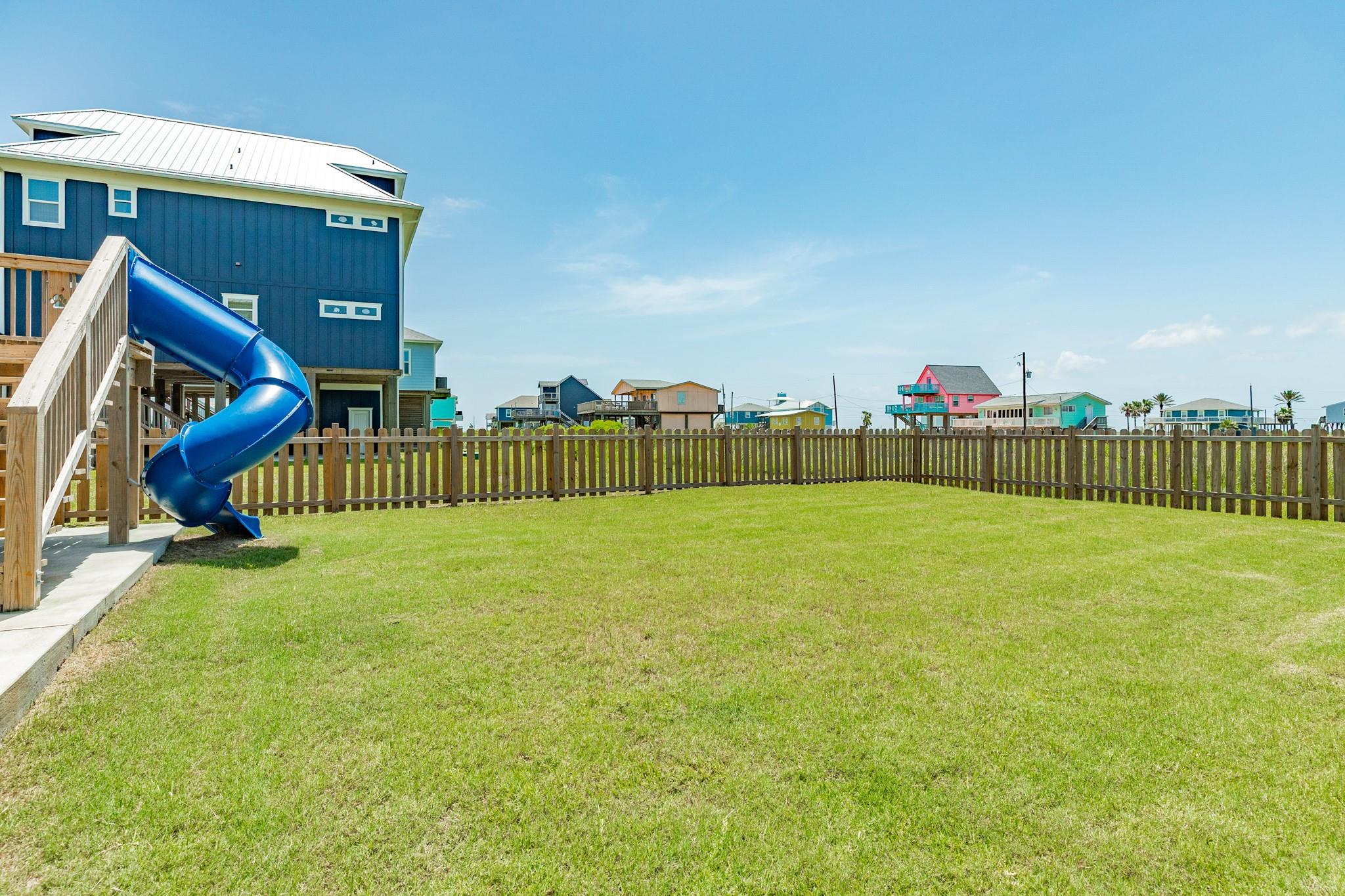Fenced backyard with a broad lawn and a blue spiral slide beside an elevated coastal home.