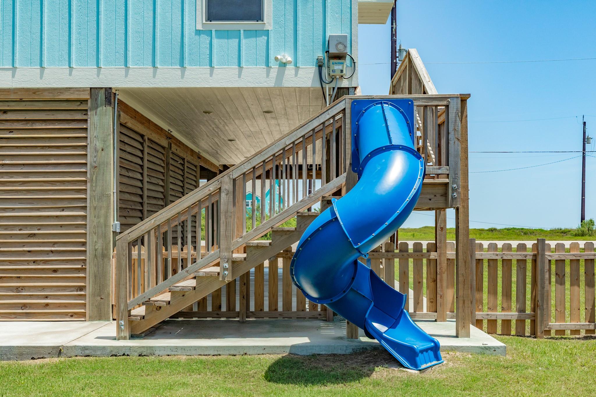 Raised wooden deck with stairs and a large blue enclosed slide beside a turquoise coastal home.