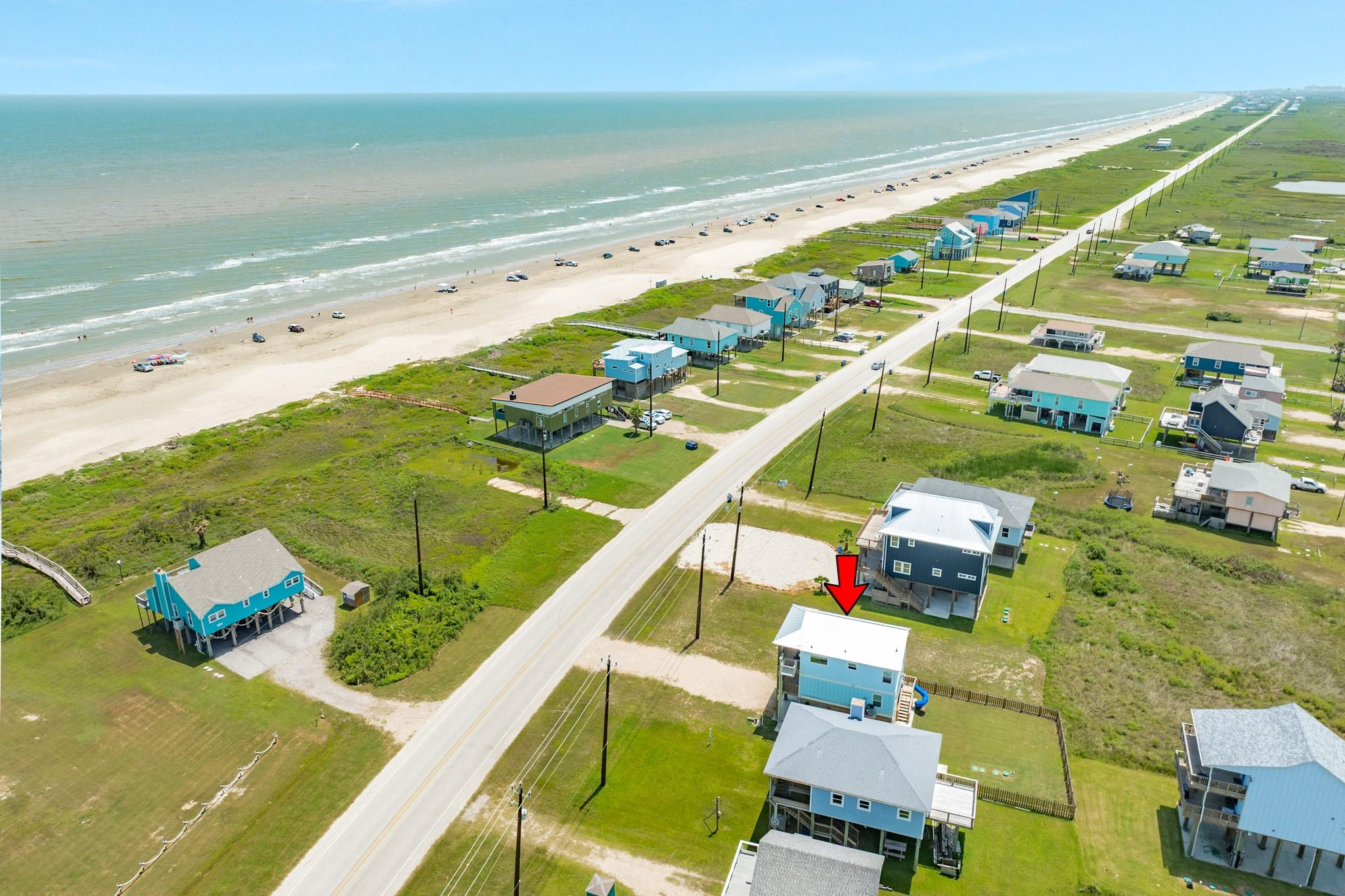 Aerial view of a light-blue raised beach house marked by an arrow near the beach and highway.