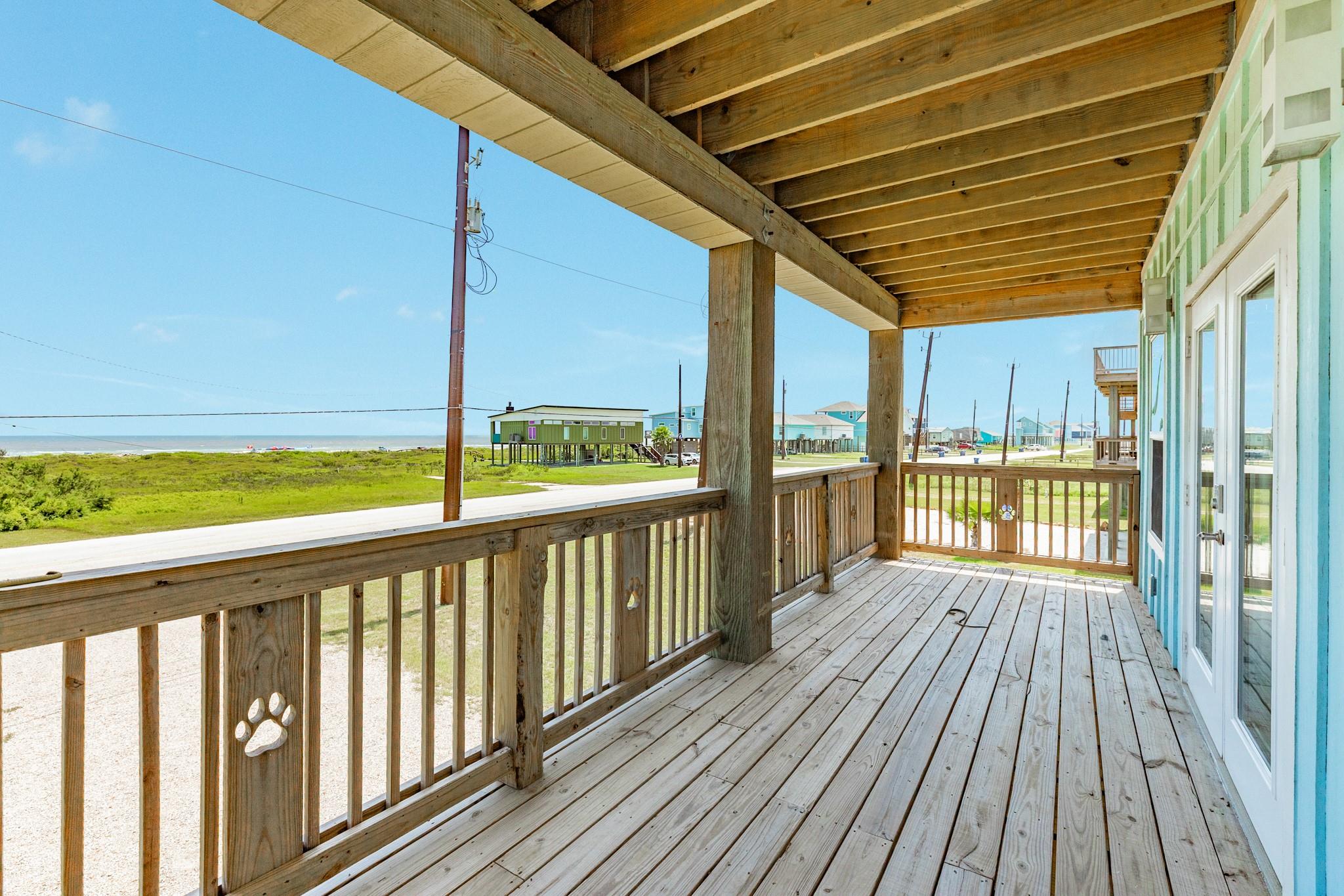Covered wooden porch overlooking coastal houses and the ocean, with a paw-print decoration on a railing post.