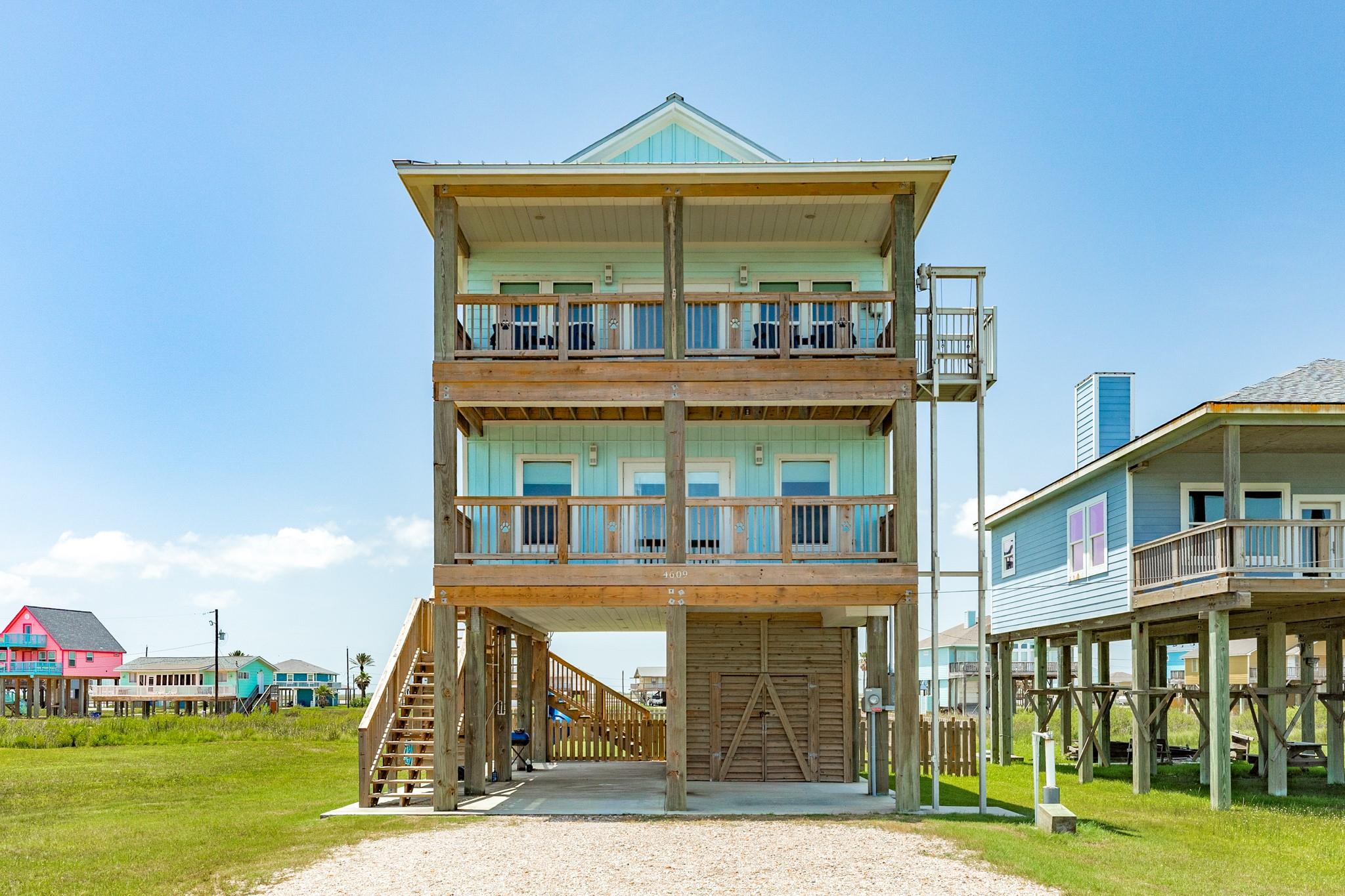 Elevated aqua coastal house with two wooden balconies and exterior stairs.