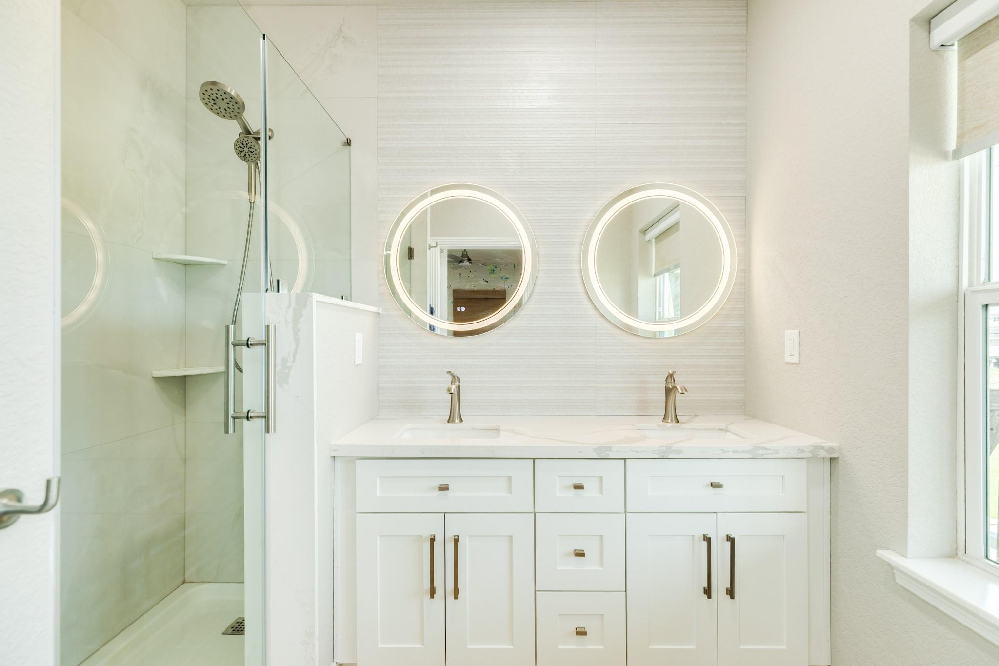 Bright bathroom with a double vanity, illuminated round mirrors, and glass-enclosed walk-in shower.
