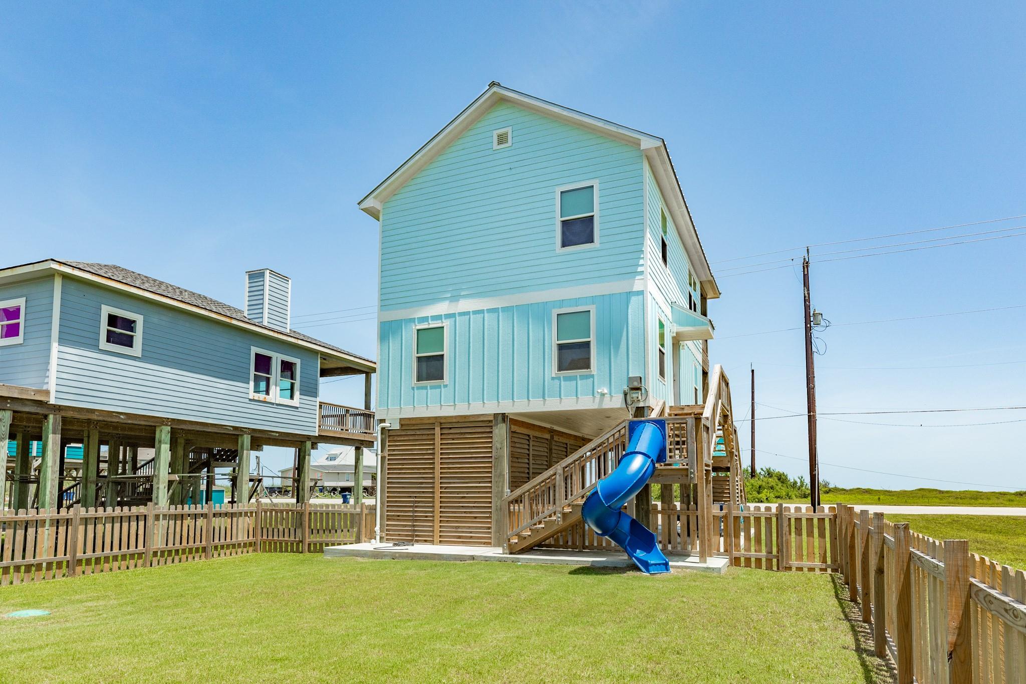 Elevated light-blue coastal house with a wooden staircase, blue spiral slide, and fenced lawn.