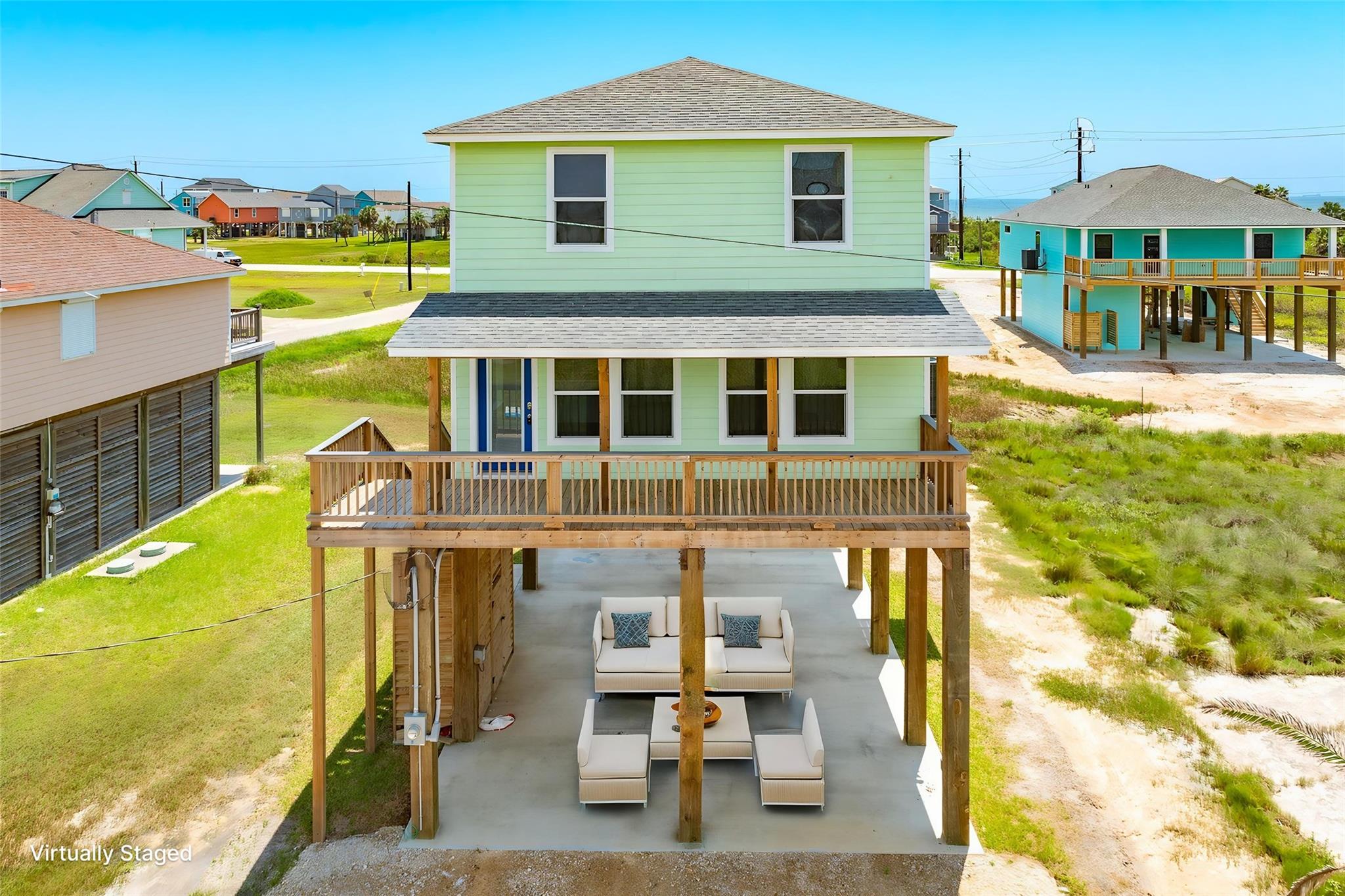Raised pale green coastal home with a wooden deck and virtually staged patio furniture.