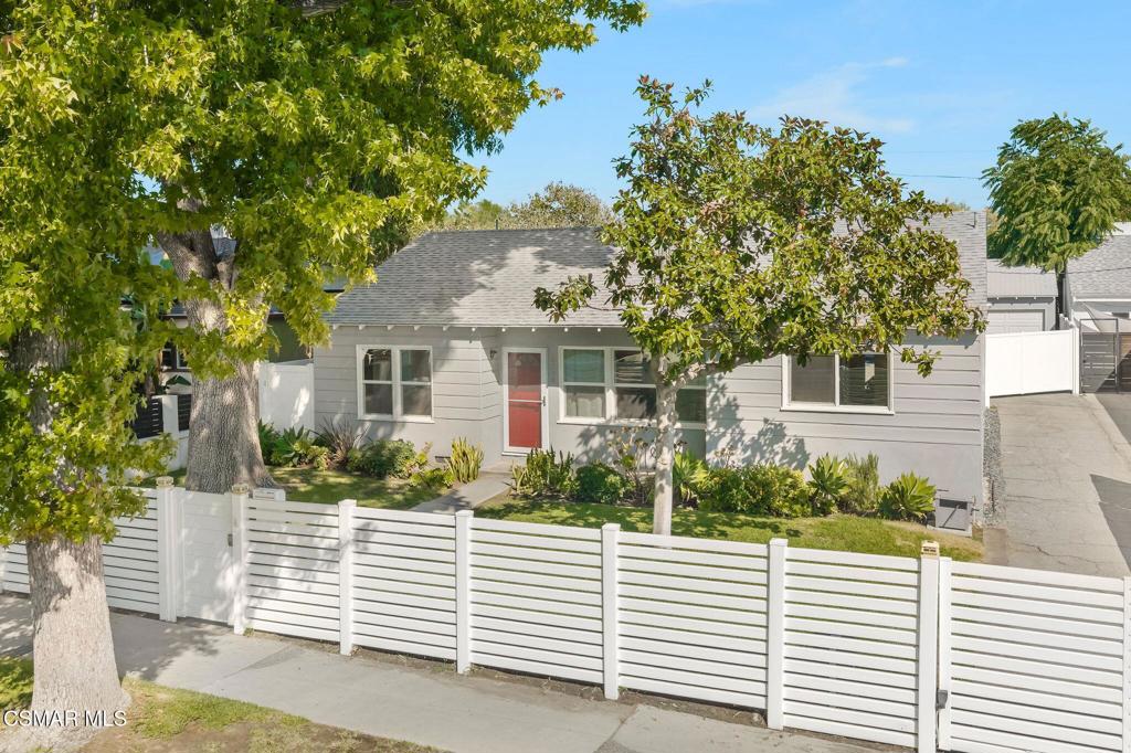 Gray single-story home with a red front door behind a white fence and mature trees.