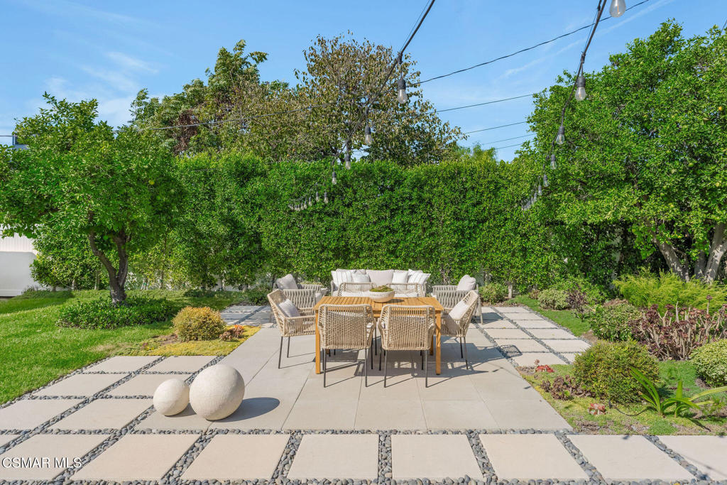 Backyard patio with wicker dining chairs, a paved terrace, mature trees, and overhead string lights.