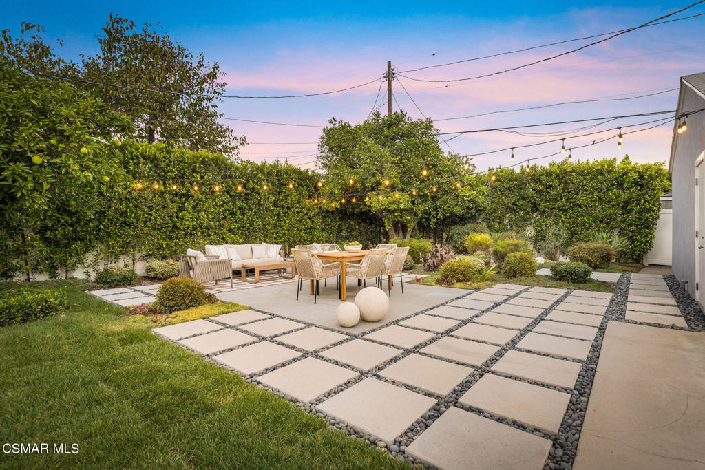 Backyard patio with stone pavers, outdoor dining furniture, and glowing string lights at dusk.