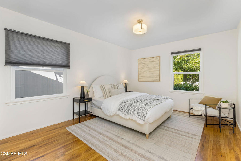 Staged bedroom with a white bed, wood floors, and a reading chair beside a garden-facing window.
