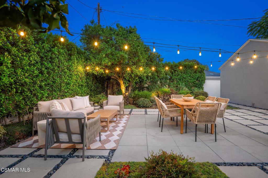 Backyard patio with dining and lounge seating beneath warm string lights at dusk.