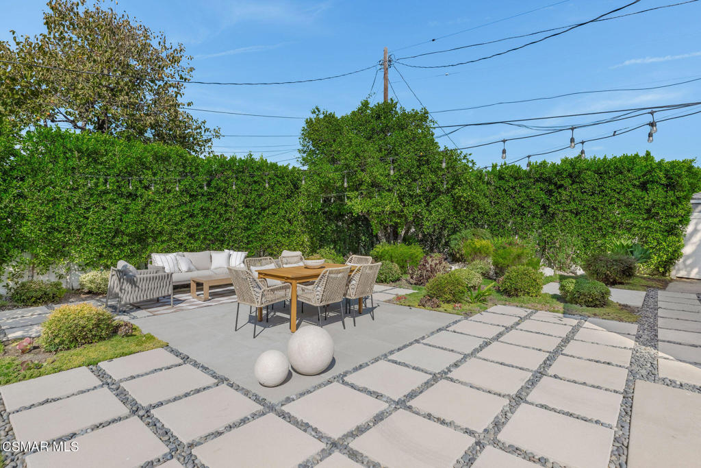Backyard patio with stone paving, outdoor dining set, lounge furniture, and a tall green privacy hedge.