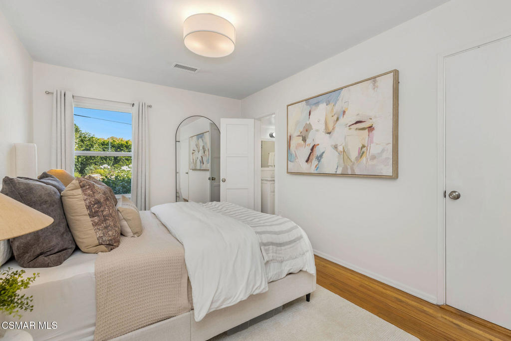 Staged bedroom with a white bed, neutral pillows, abstract wall art, and a window overlooking trees.