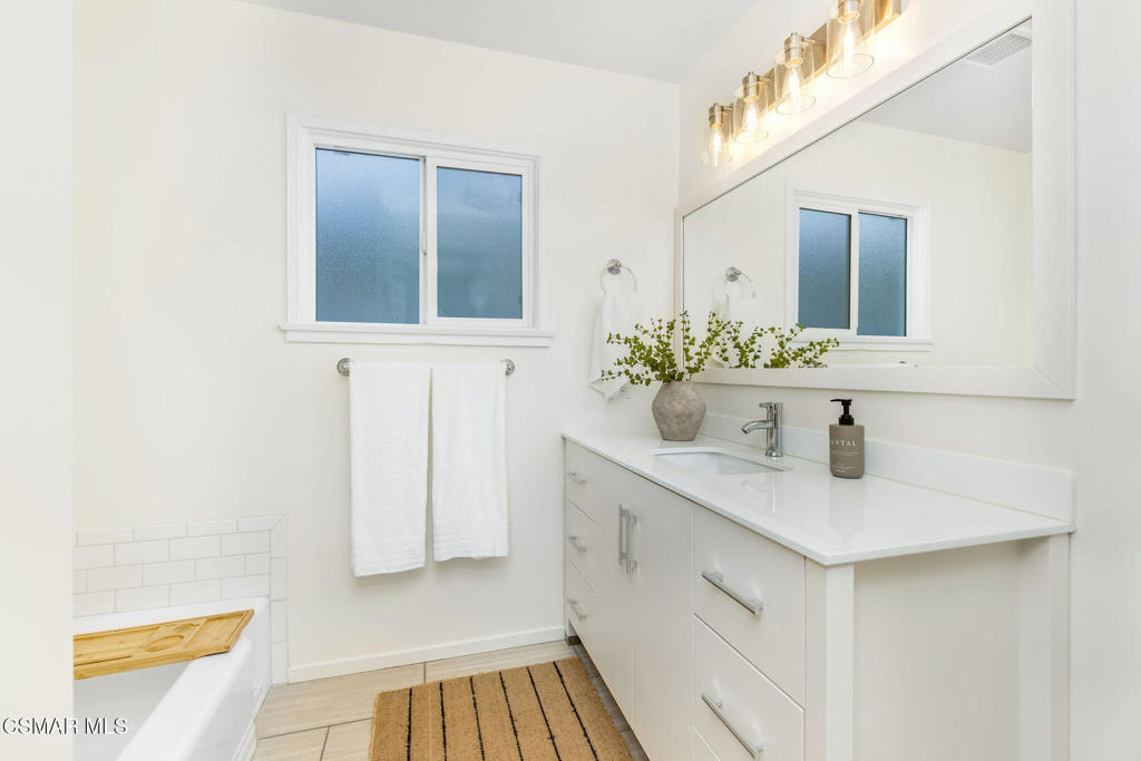 Bright bathroom with a white vanity, large mirror, window, and bathtub with a wooden bath tray.