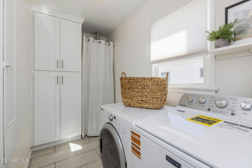 Laundry room with white washer and dryer beneath a window, beside tall white storage cabinets.