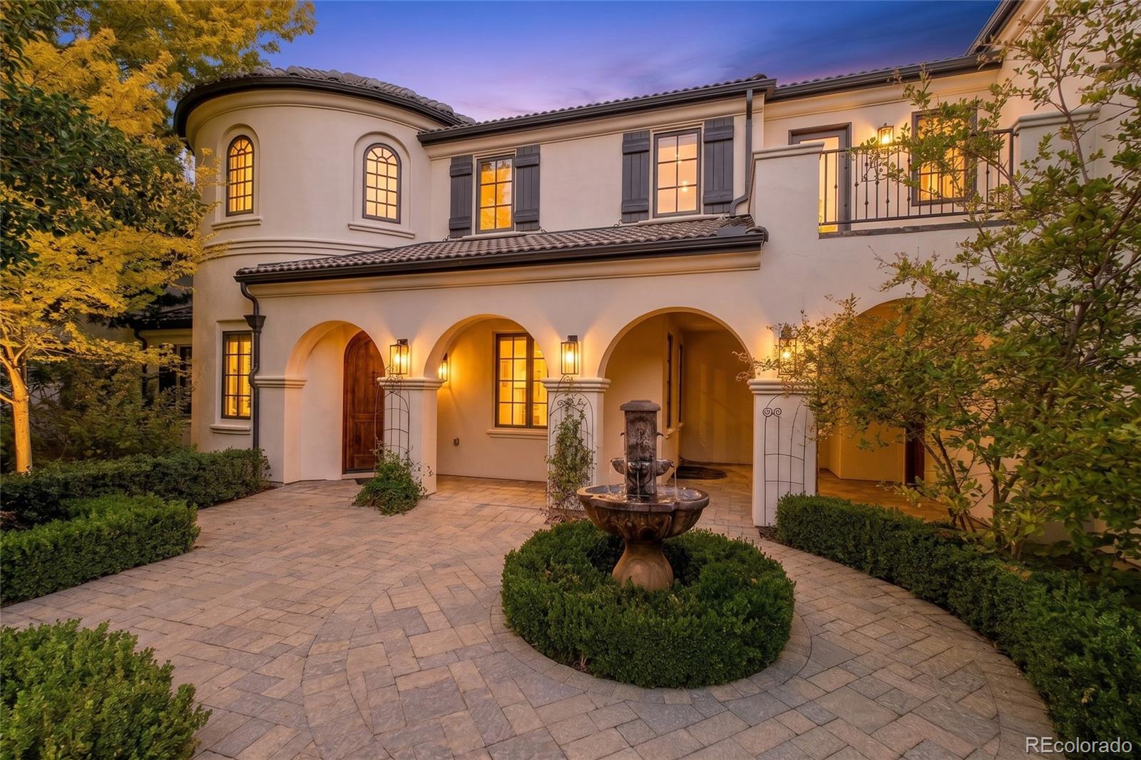 Mediterranean-style home with arched entryways and a fountain in its paved courtyard at dusk.