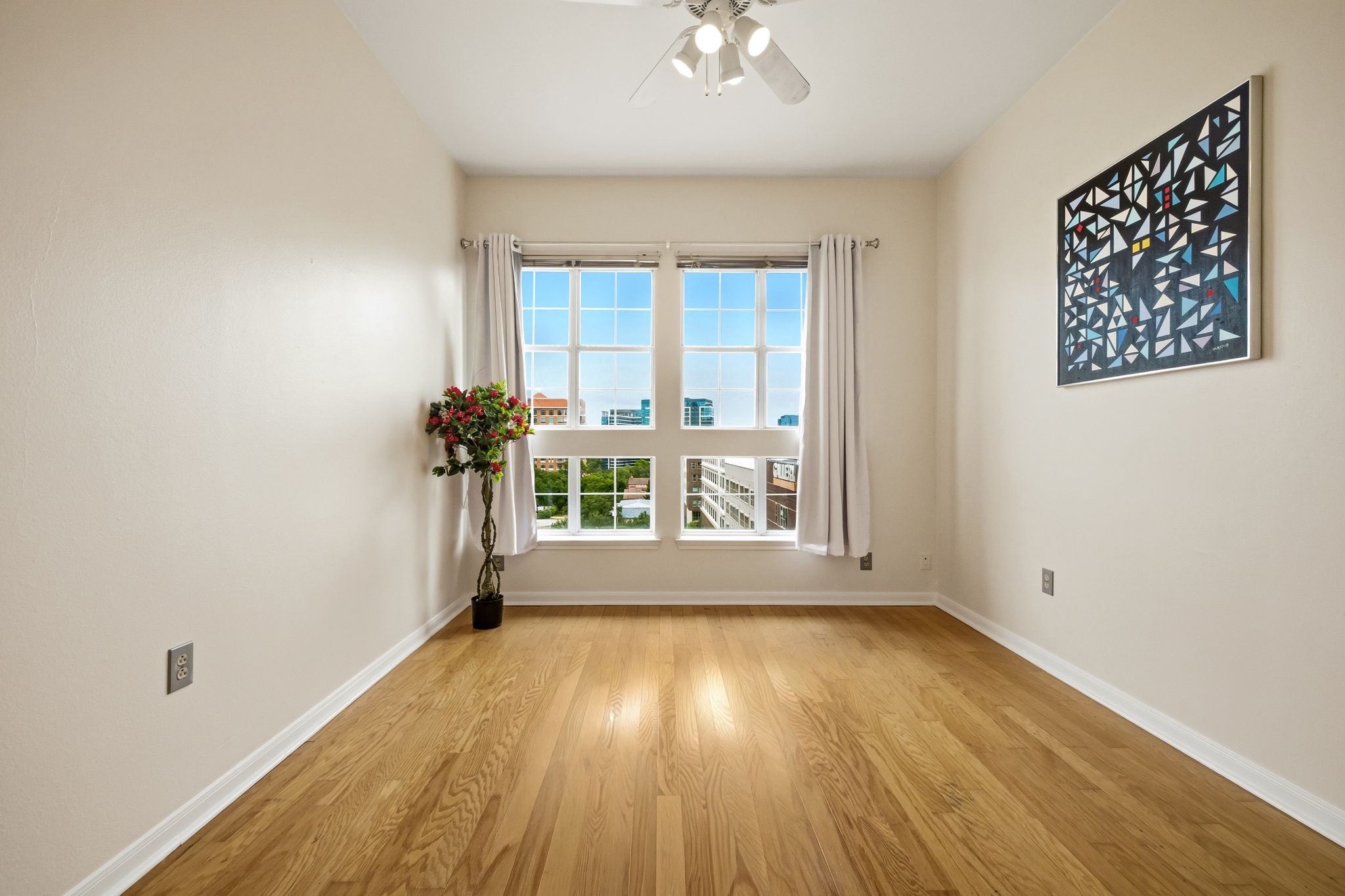 Bright empty room with hardwood floors, large windows, a flowering plant, and geometric wall art.