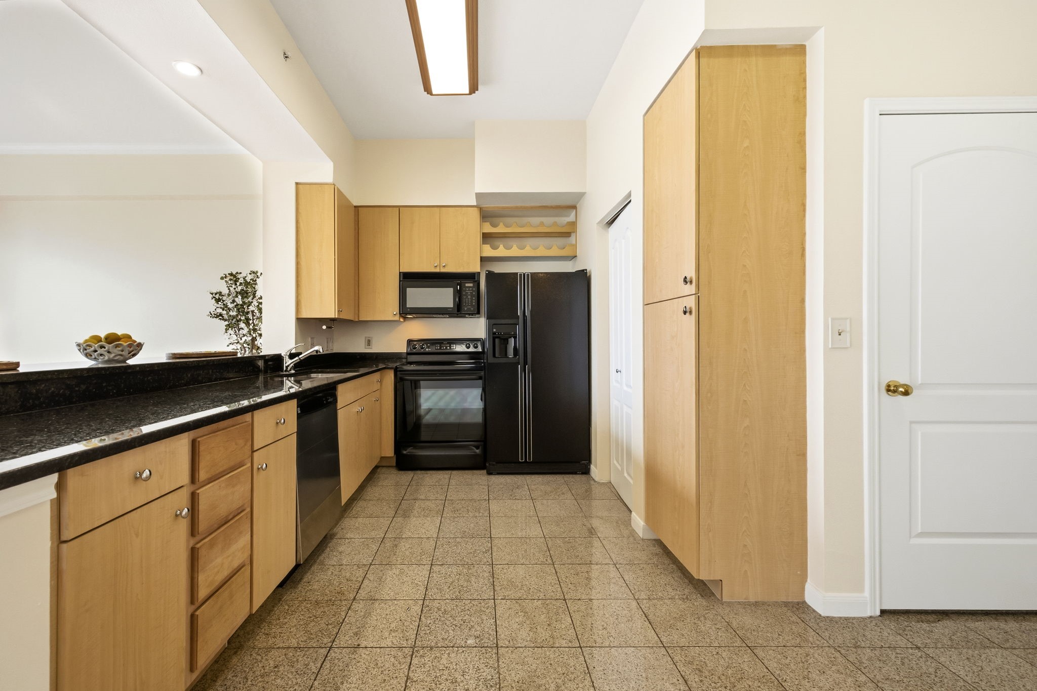 Galley kitchen with light wood cabinets, dark countertops, and black appliances.