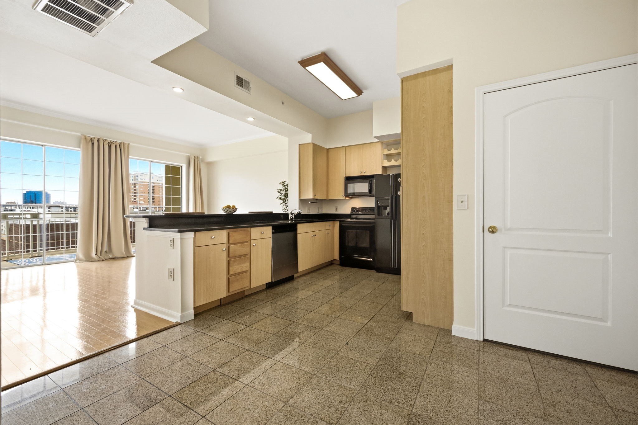 Condo kitchen with light wood cabinets and black counters, opening to a bright living area with city views.