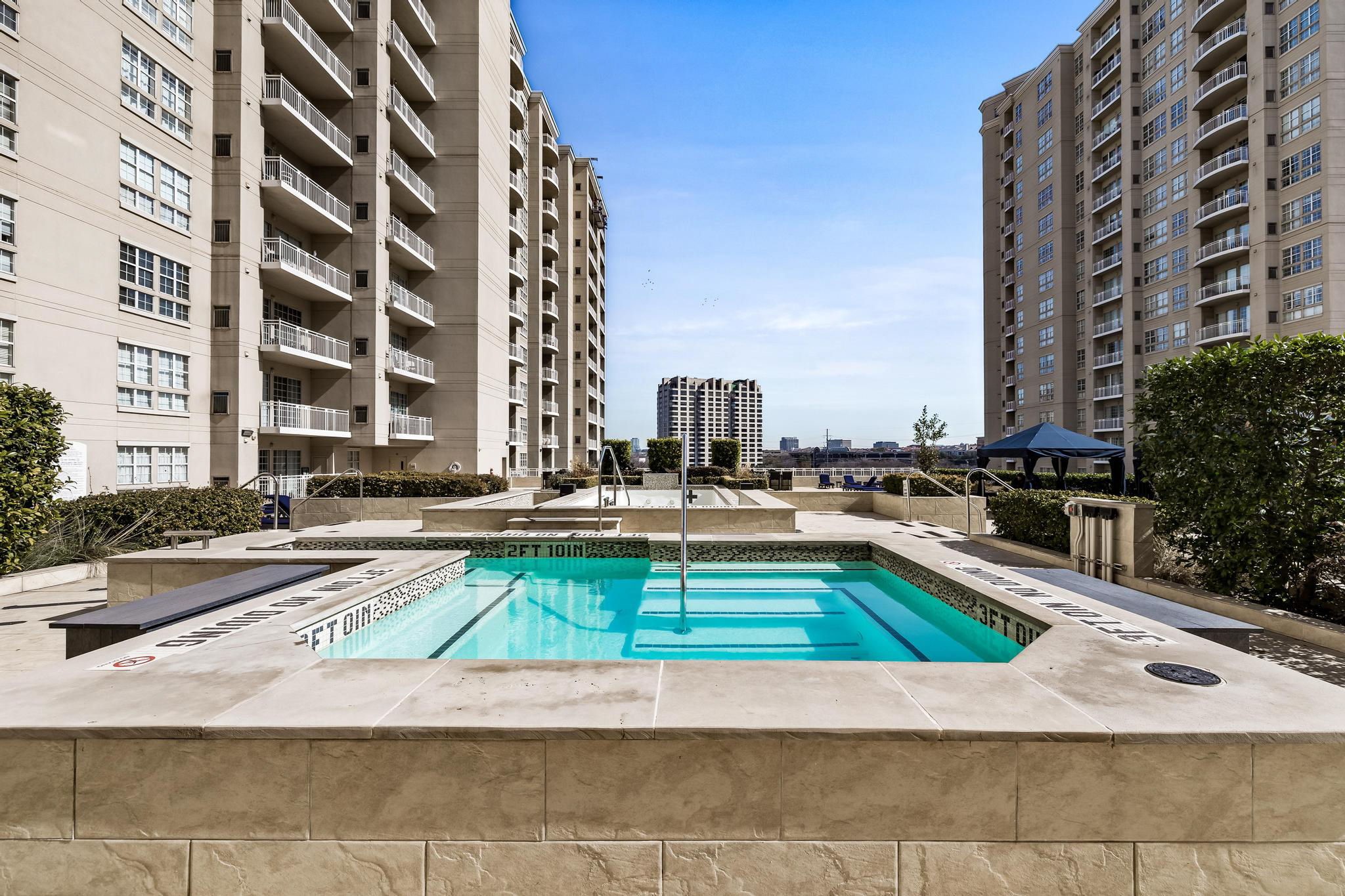 Outdoor pool and raised spa between high-rise buildings, with a city skyline in the distance.