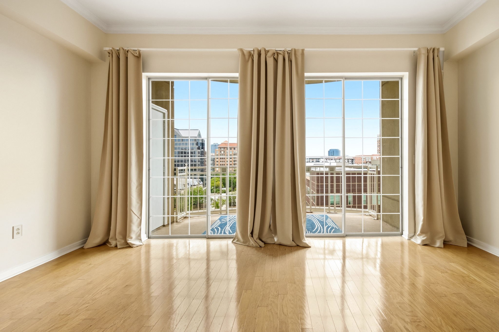 Empty condo room with hardwood floors and tall curtained glass doors opening to a balcony and city view.