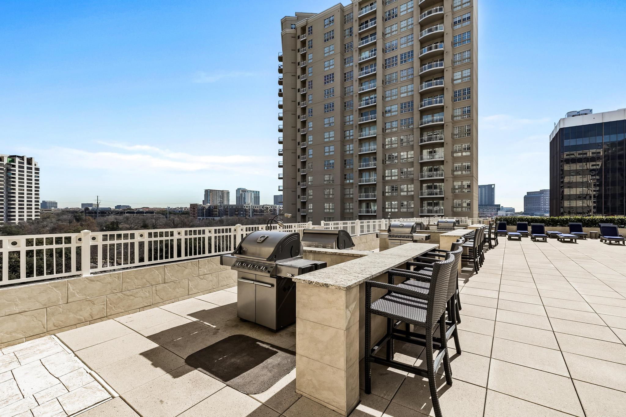 Rooftop terrace with outdoor grills, bar seating, lounge chairs, and a Dallas skyline view.