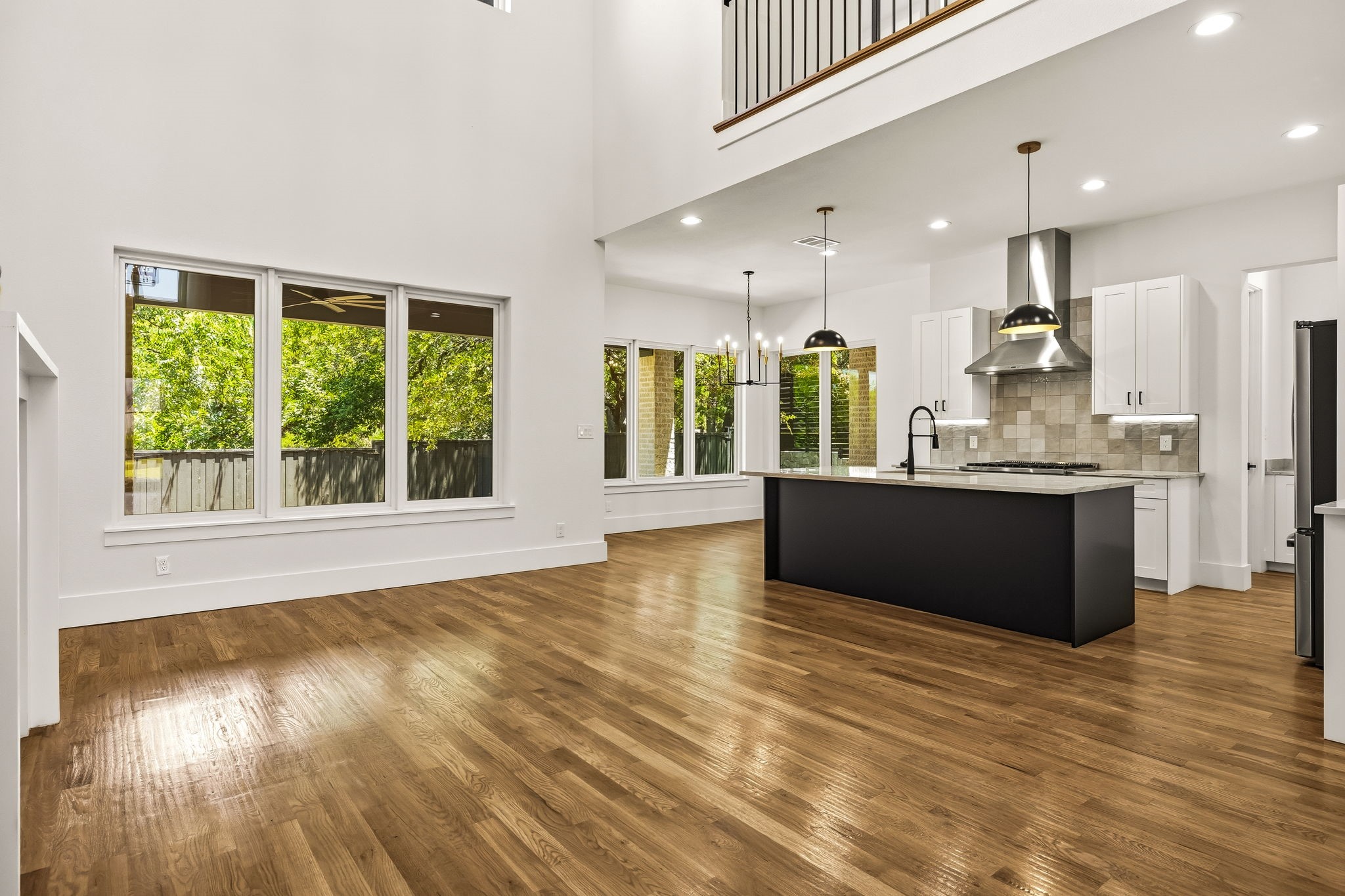 Open-plan kitchen with a large black island, hardwood floors, tall windows, and an upper-level railing.