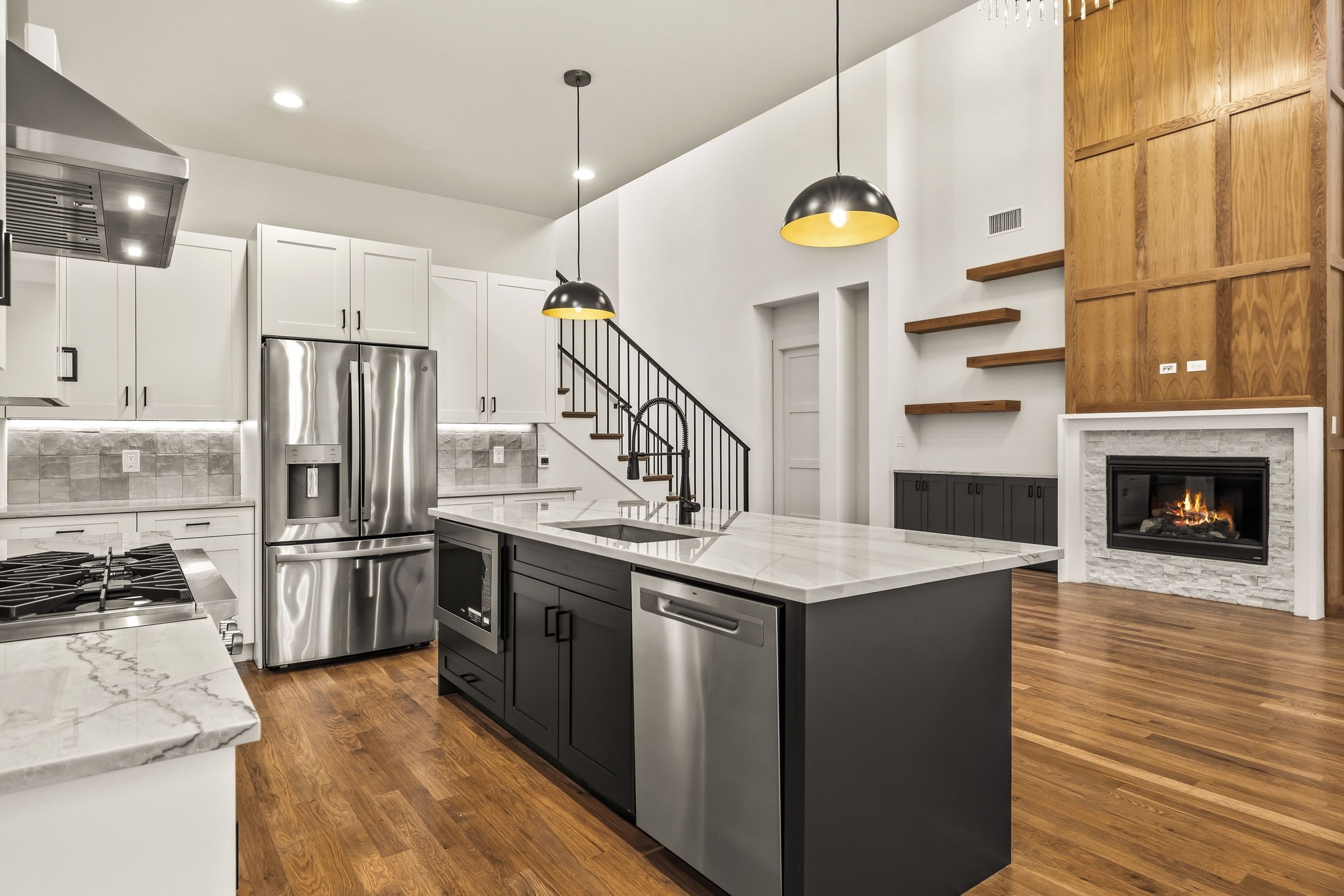 Open-plan kitchen with a large dark island, stainless appliances, and a fireplace against a wood feature wall.