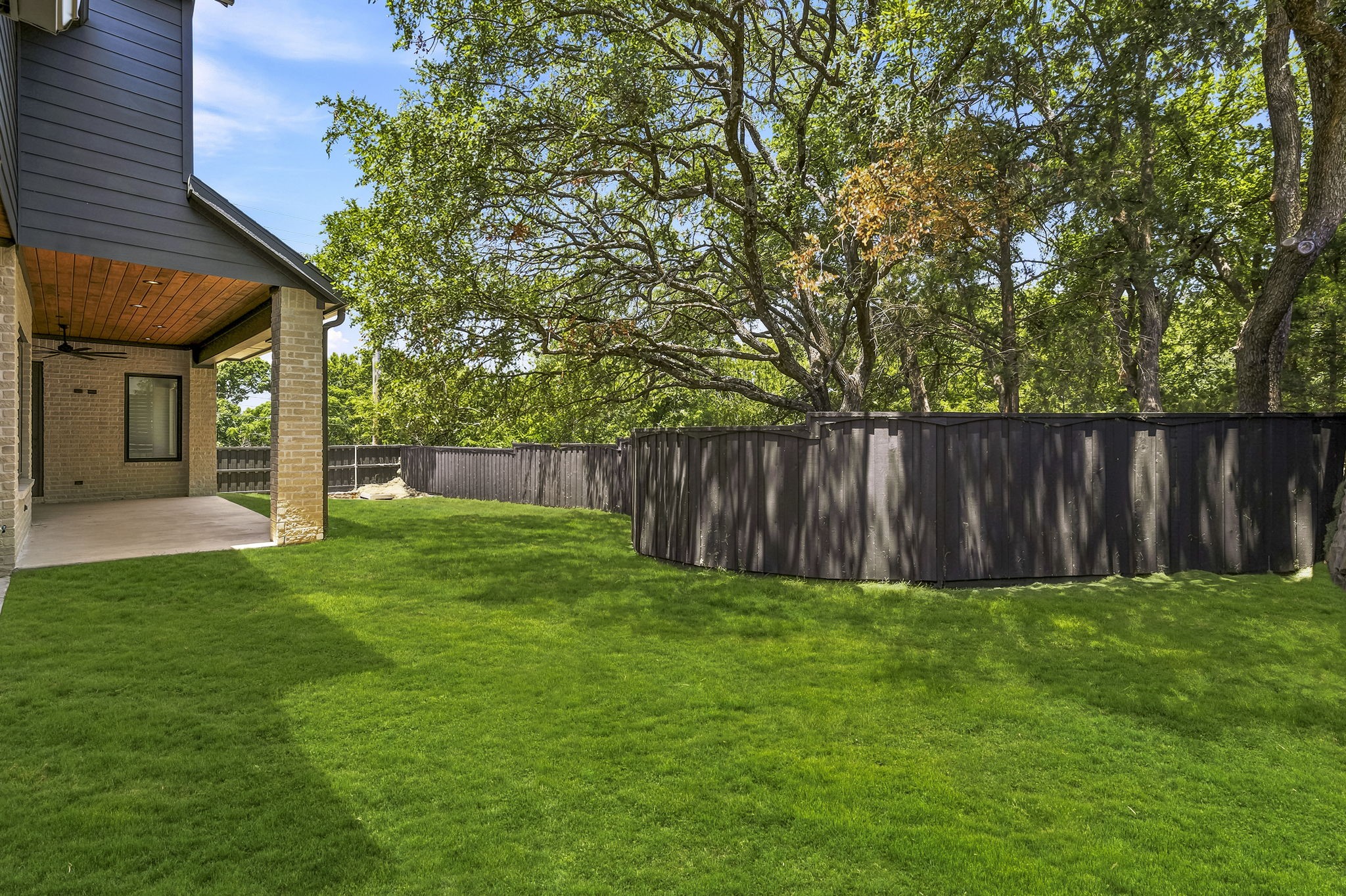 Backyard with a covered stone patio, broad lawn, mature trees, and dark privacy fence