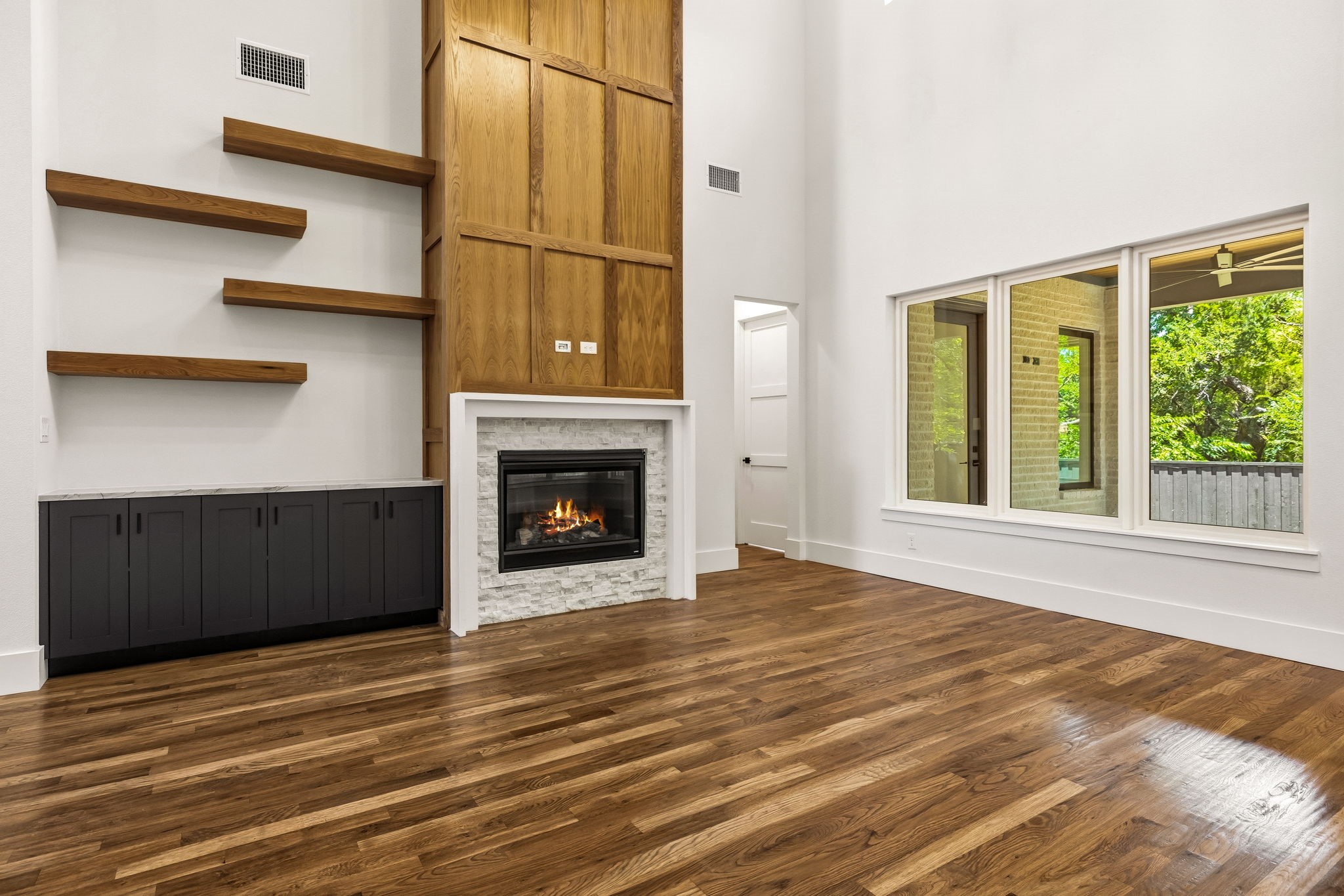 Unfurnished living room with a stone fireplace, tall wood paneling, floating shelves, and large windows.