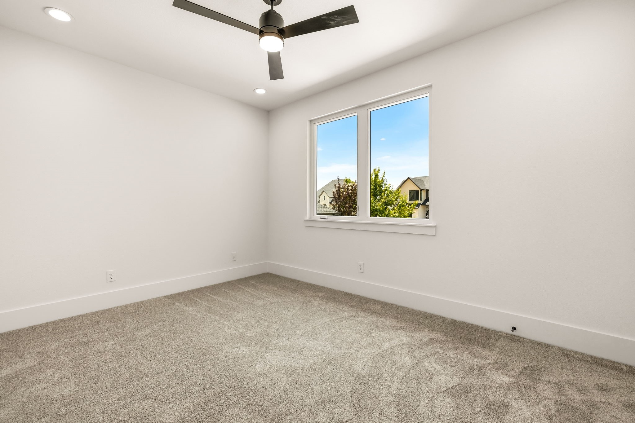 Empty carpeted room with a ceiling fan and large window overlooking neighboring homes.