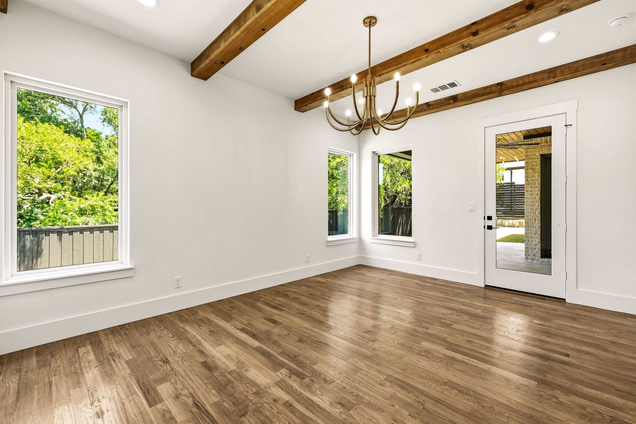 Empty room with exposed wood ceiling beams, a brass chandelier, and windows overlooking leafy trees.