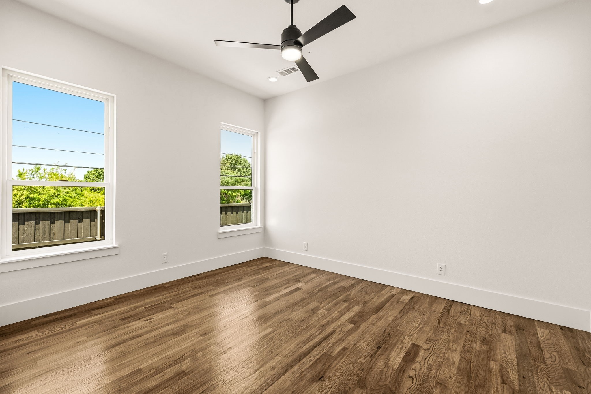 Empty room with wood plank flooring, two windows, and a black ceiling fan.