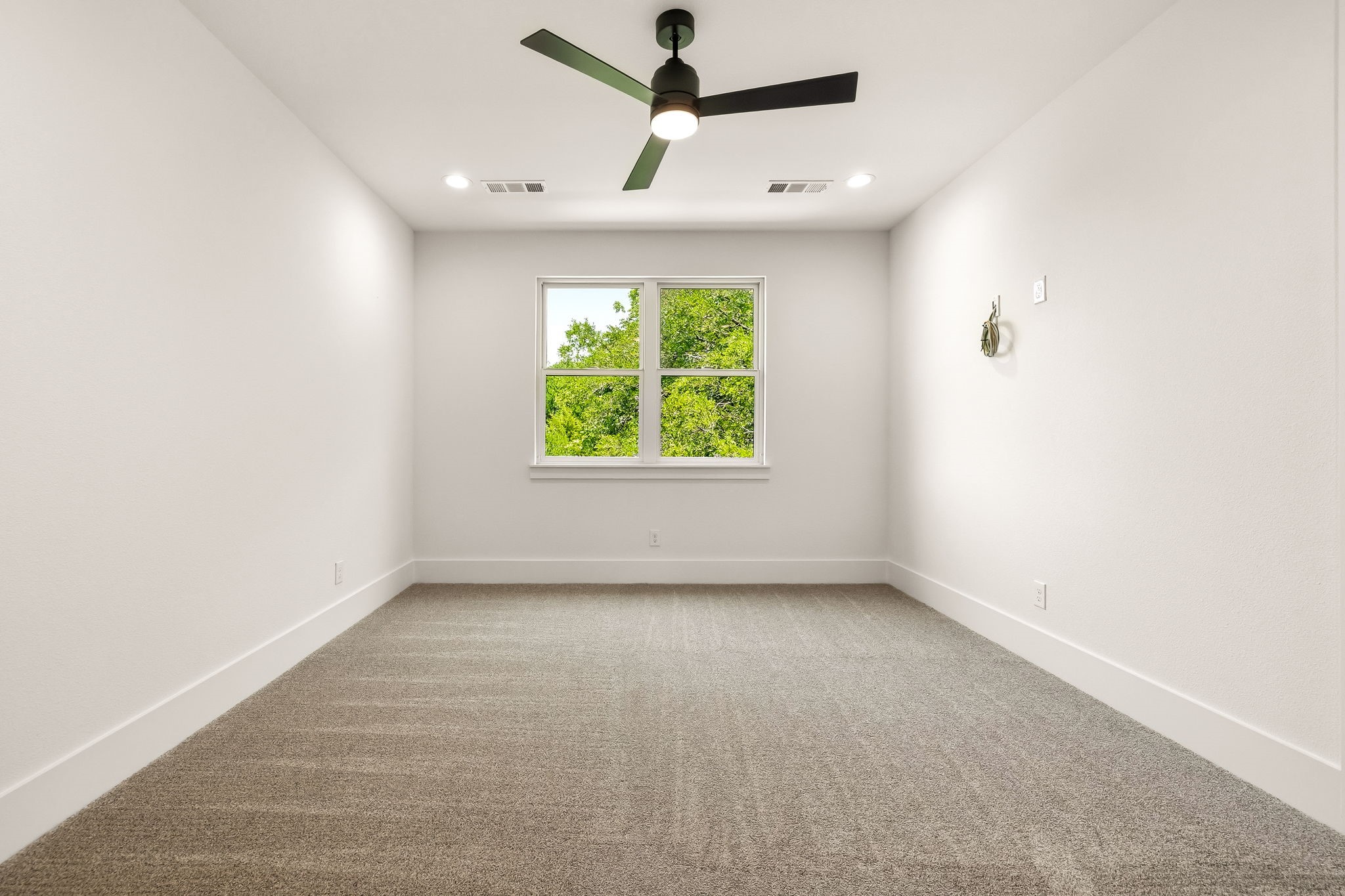Empty carpeted room with a ceiling fan and a window overlooking leafy trees.
