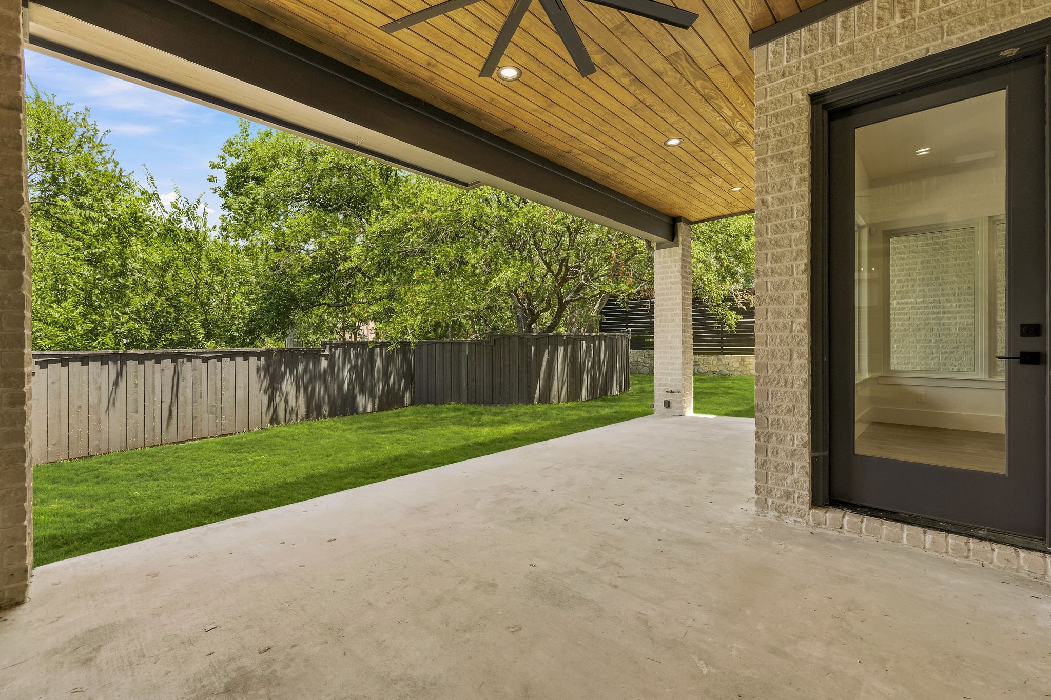 Covered patio with a wood ceiling and fan overlooking a fenced lawn and trees.