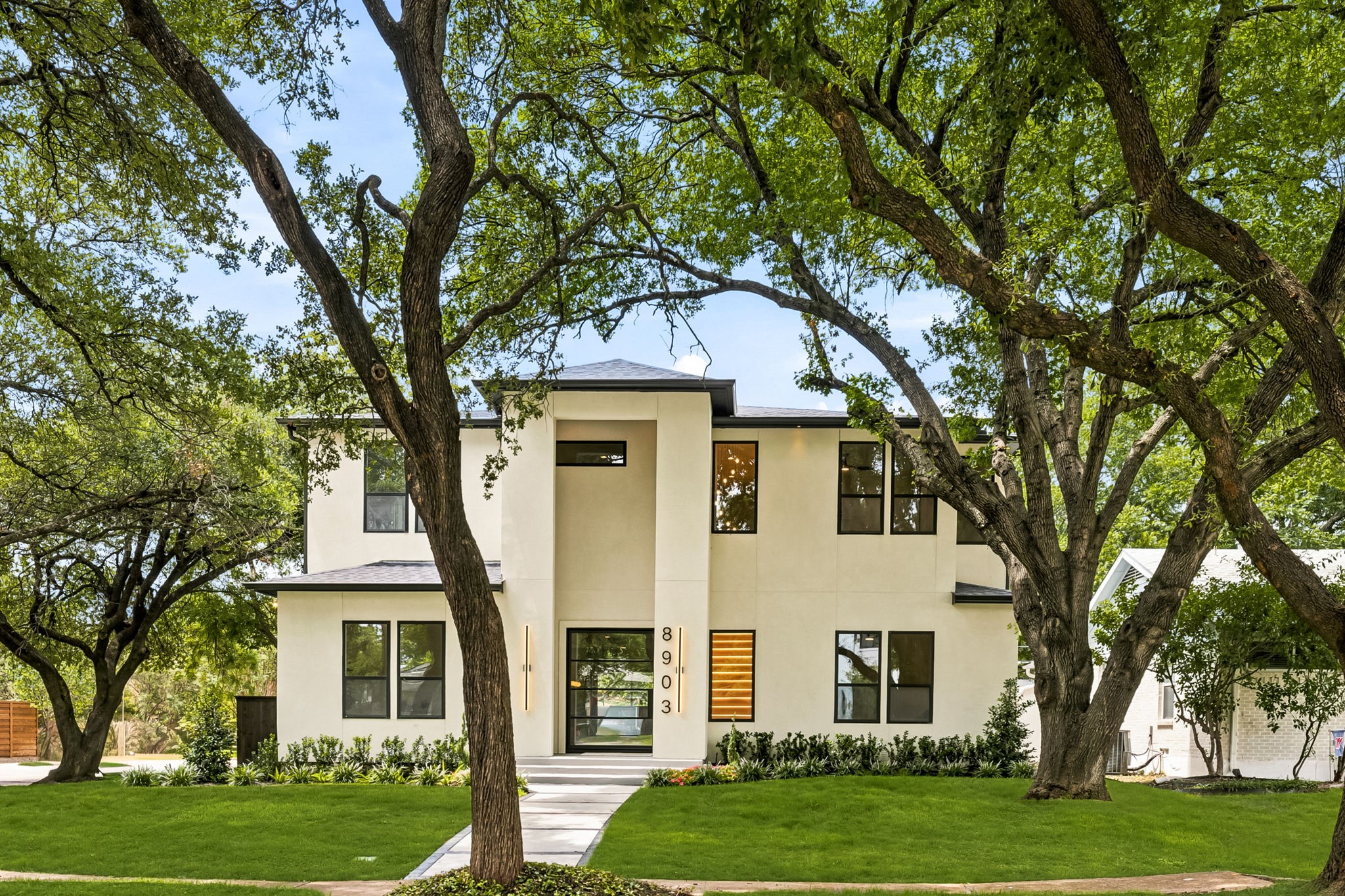 Modern two-story home with a cream facade, black-framed windows, and mature trees around the front lawn.