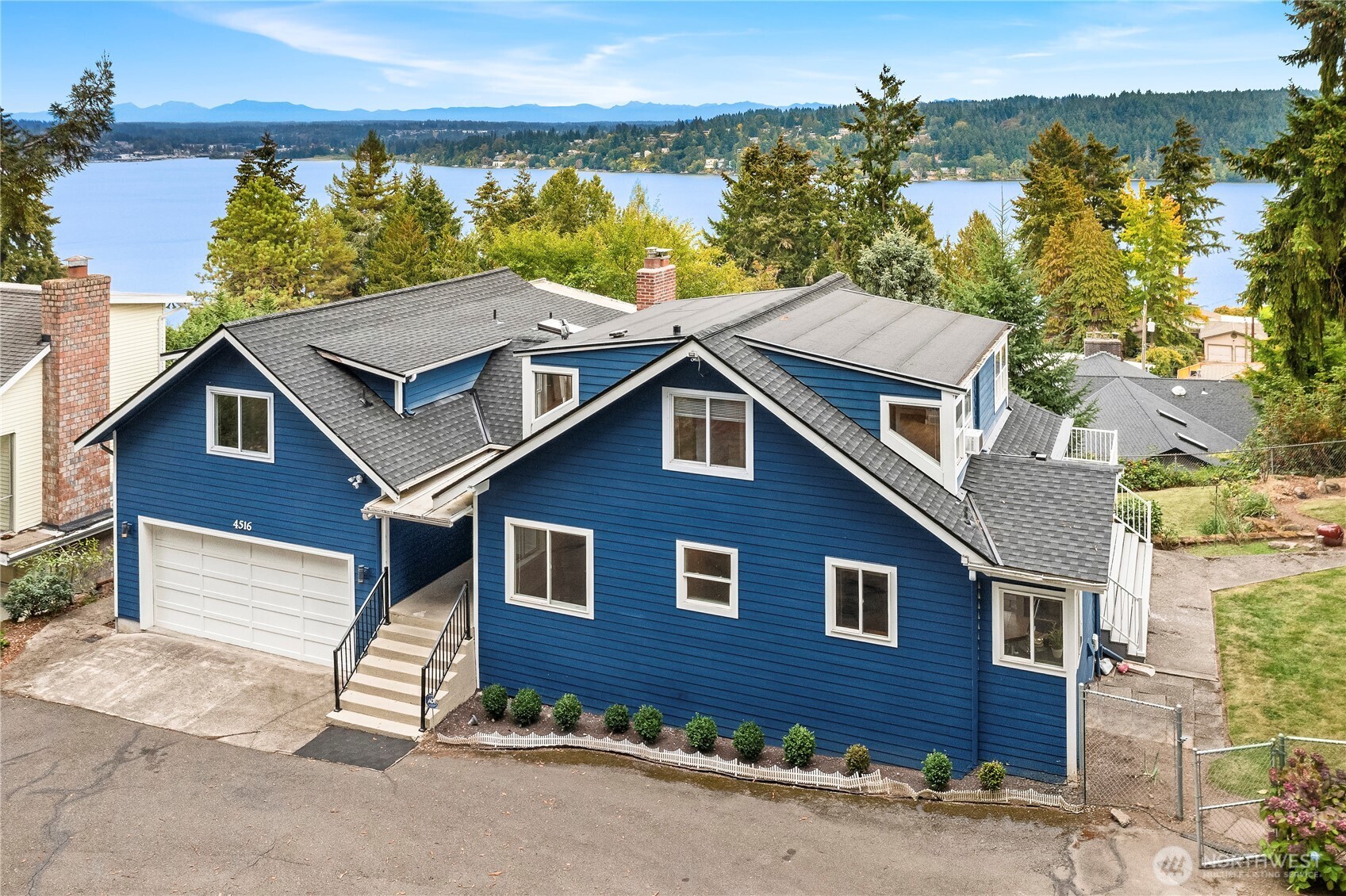 Blue multi-level home with a two-car garage, overlooking a lake and distant mountains.