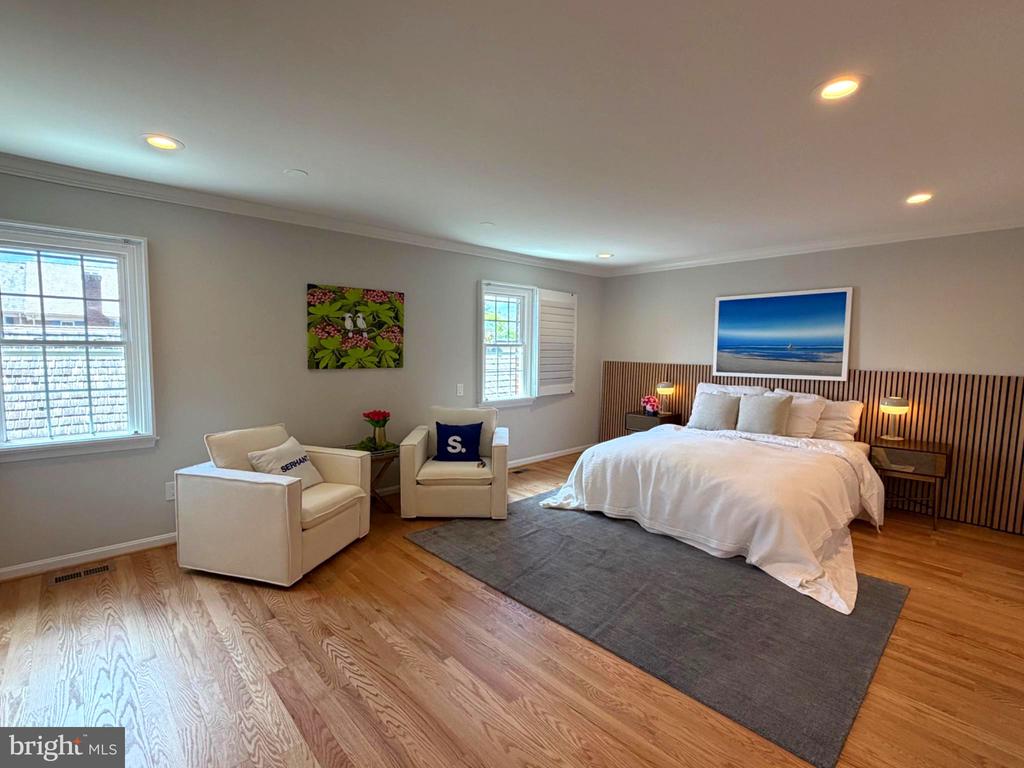 Staged bedroom with a white bed, slatted wood headboard, cream armchairs, and hardwood floors.