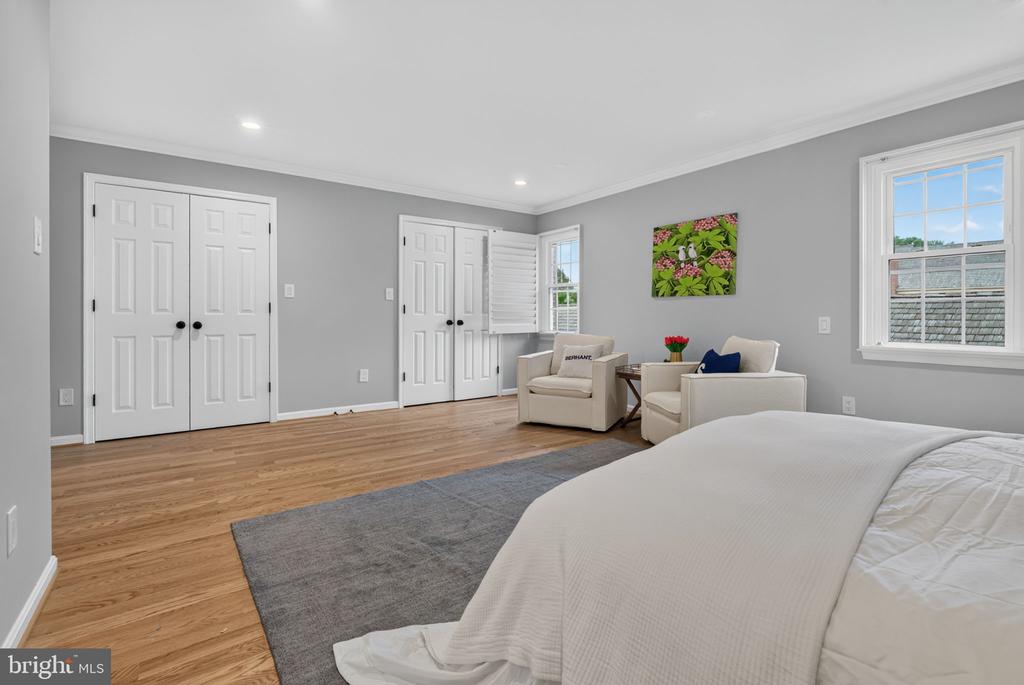Staged bedroom with a large bed, gray rug, light wood floors, and a sitting area by the window.