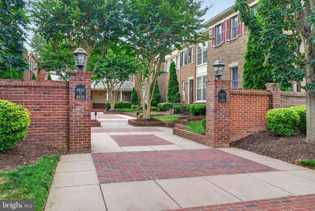 Brick townhome community entrance with lantern-topped gateposts and a tree-lined paved walkway.