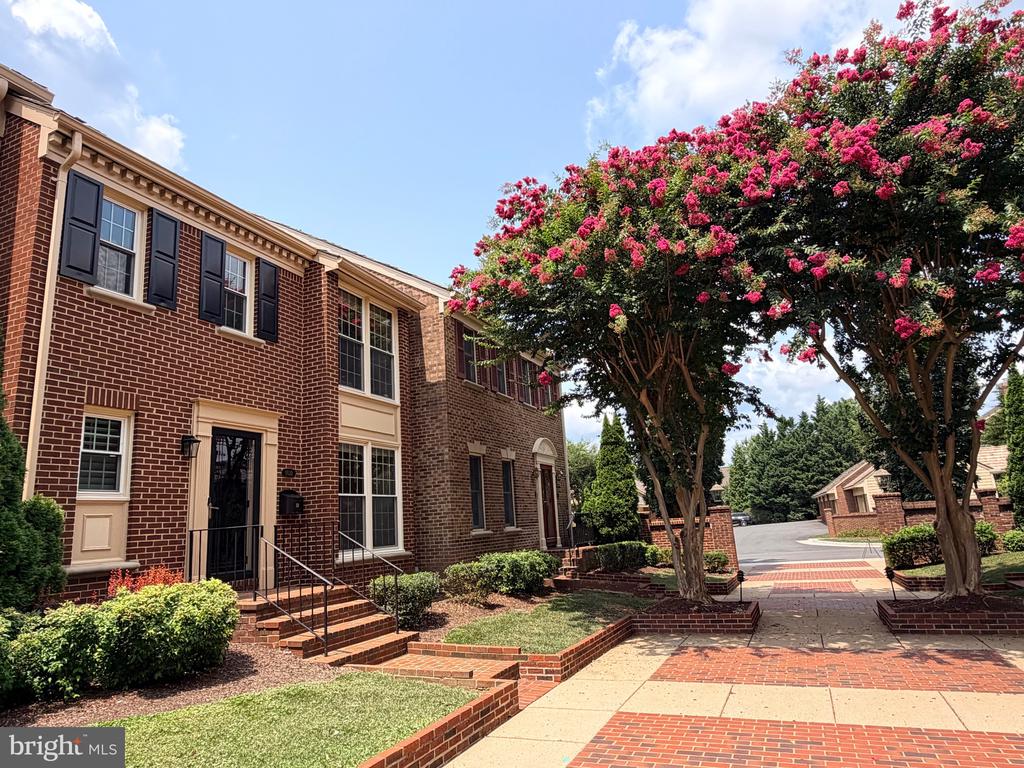 Brick townhomes with black shutters beside a pink-flowering tree and landscaped walkway.