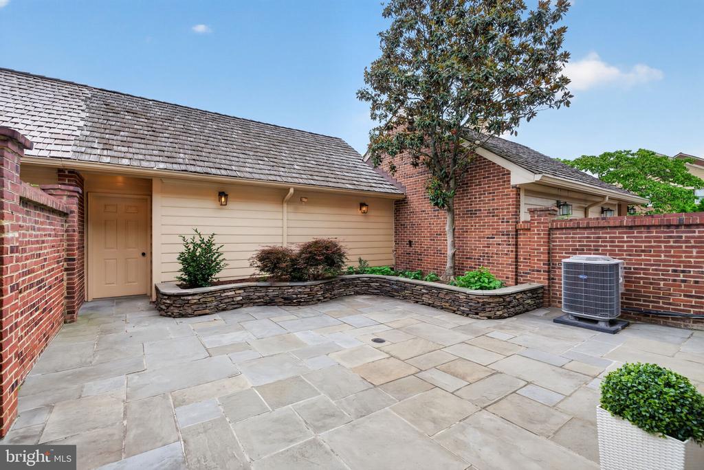 Stone-paved courtyard with a beige entrance, brick walls, raised planting beds, and a tree.