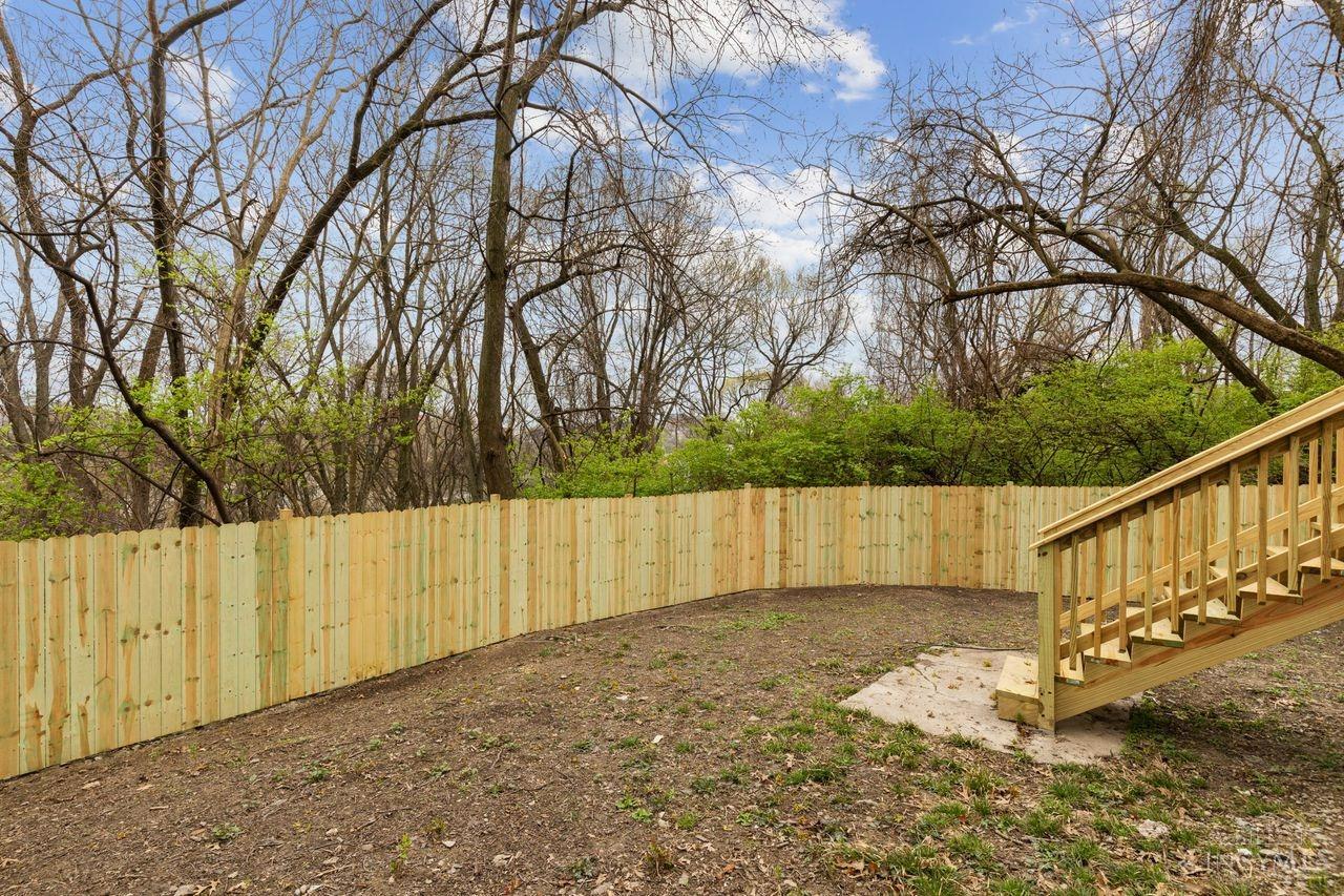 Fenced backyard with a wooden staircase, bare trees, and green shrubs beyond the fence.