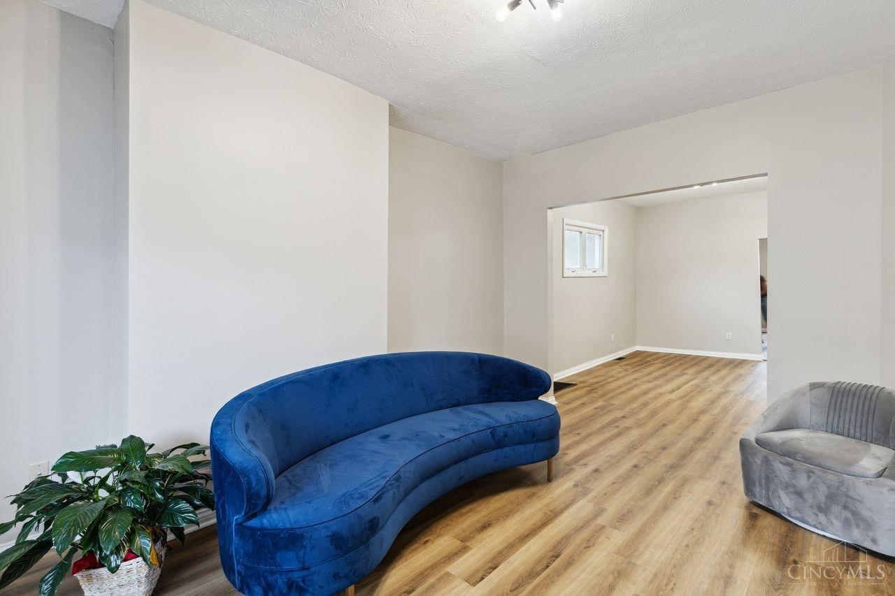 Living room with a curved blue sofa, gray chair, and wood-look floors opening to a bright adjoining room.