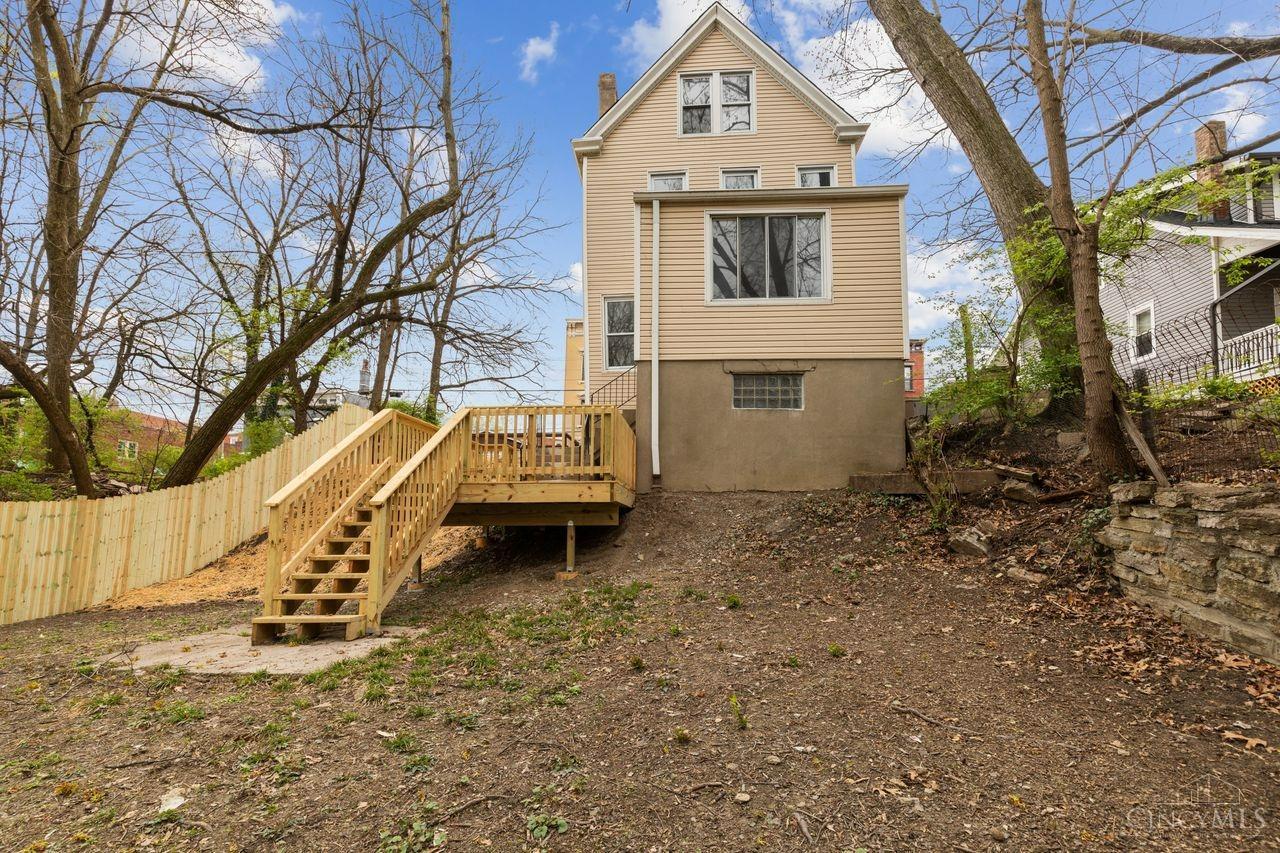 Rear of a beige multi-story house with a raised wooden deck and stairs in a sloping, fenced yard.